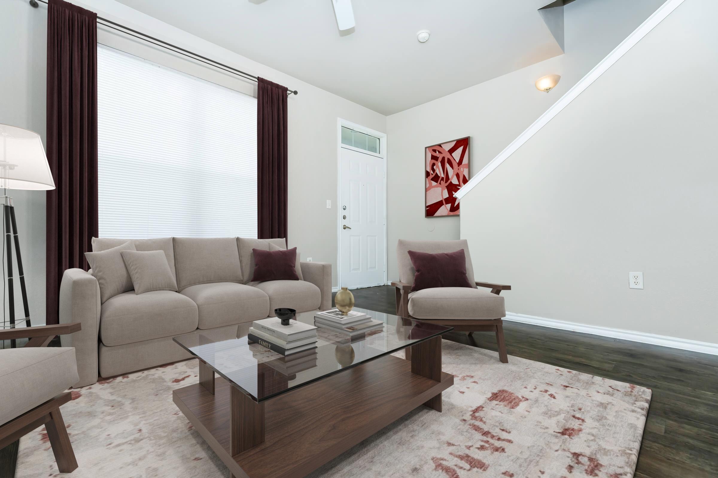 A modern living room featuring a beige sofa with decorative pillows, two armchairs, and a glass coffee table. The walls are painted light, with a geometric artwork on display. Natural light streams through a large window with sheer curtains, and a staircase is visible in the background.