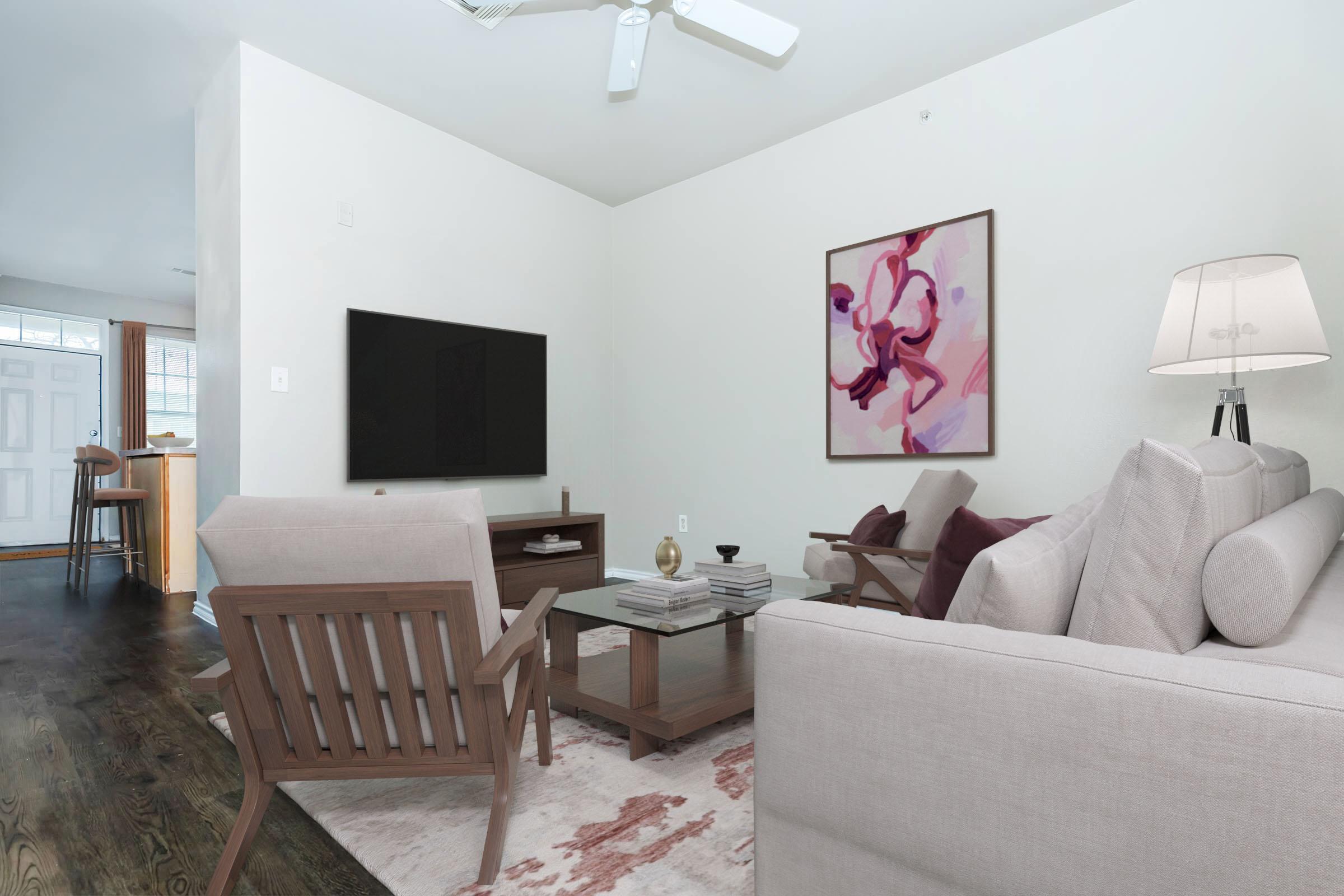 A cozy living room featuring a light-colored sofa and wooden armchairs. A modern television is mounted on the wall opposite a stylish coffee table. Decor includes a colorful abstract painting and a table lamp. The space is well-lit, with a view of an entrance in the background.