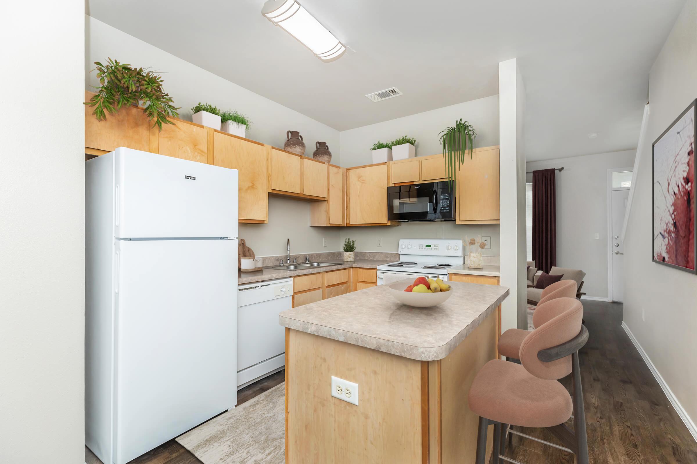Modern kitchen featuring light wood cabinetry, a white refrigerator, a sink, and a microwave. A countertop island with bar stools is in the foreground, and there are decorative plants on the shelves. Natural light illuminates the space, creating a bright and inviting atmosphere.