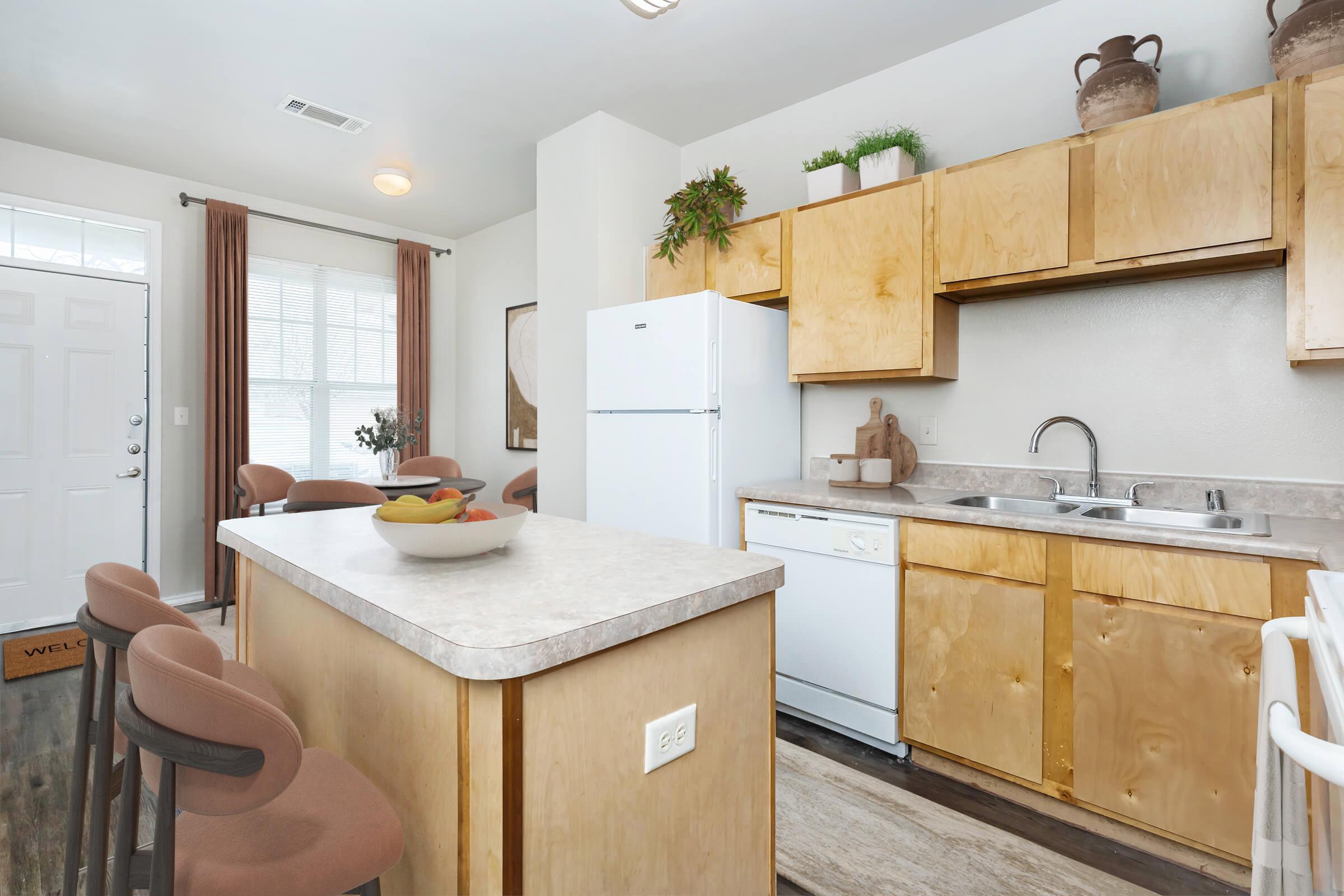 A modern kitchen featuring light wood cabinetry, a white refrigerator, and a dishwasher. The countertop includes a bowl of fruit and is accompanied by barstools. Natural light enters through a nearby window, with decorative plants and a welcoming atmosphere. The entry door is visible in the background.