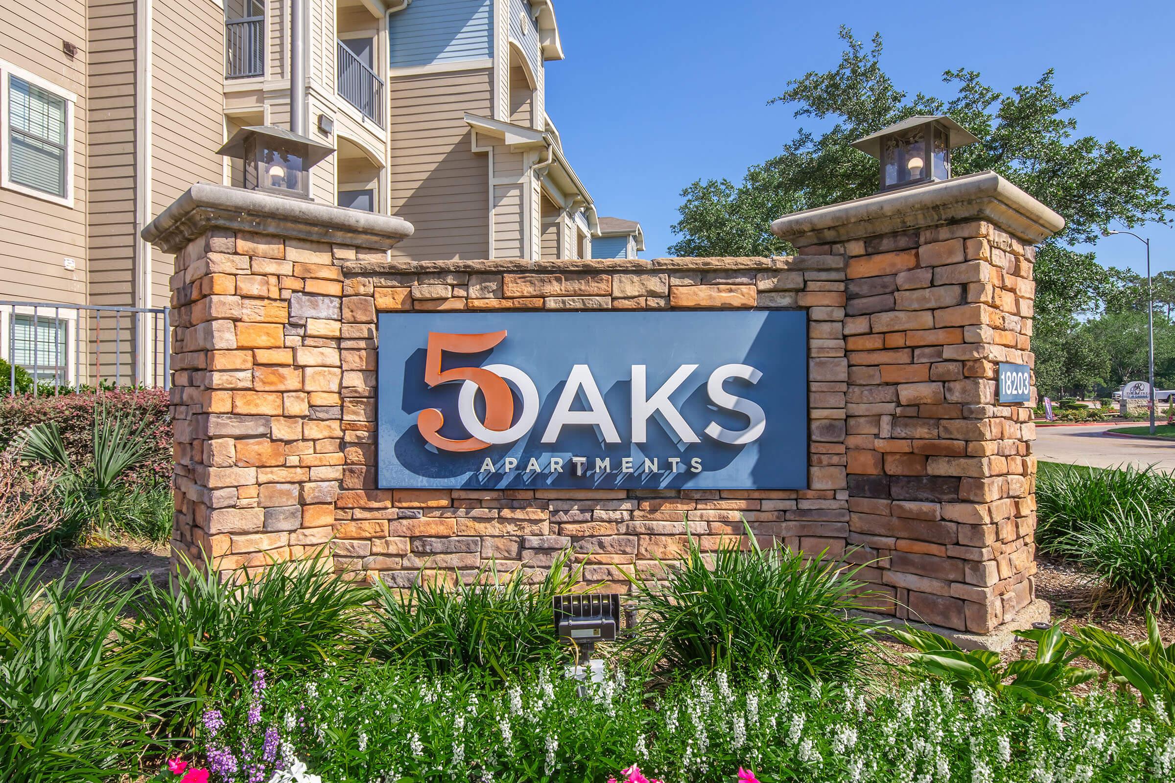 Sign for 5 Oaks Apartments featuring a modern design with a stone base and stylish lettering. The background includes well-maintained greenery and blue skies, conveying a welcoming atmosphere for potential residents.