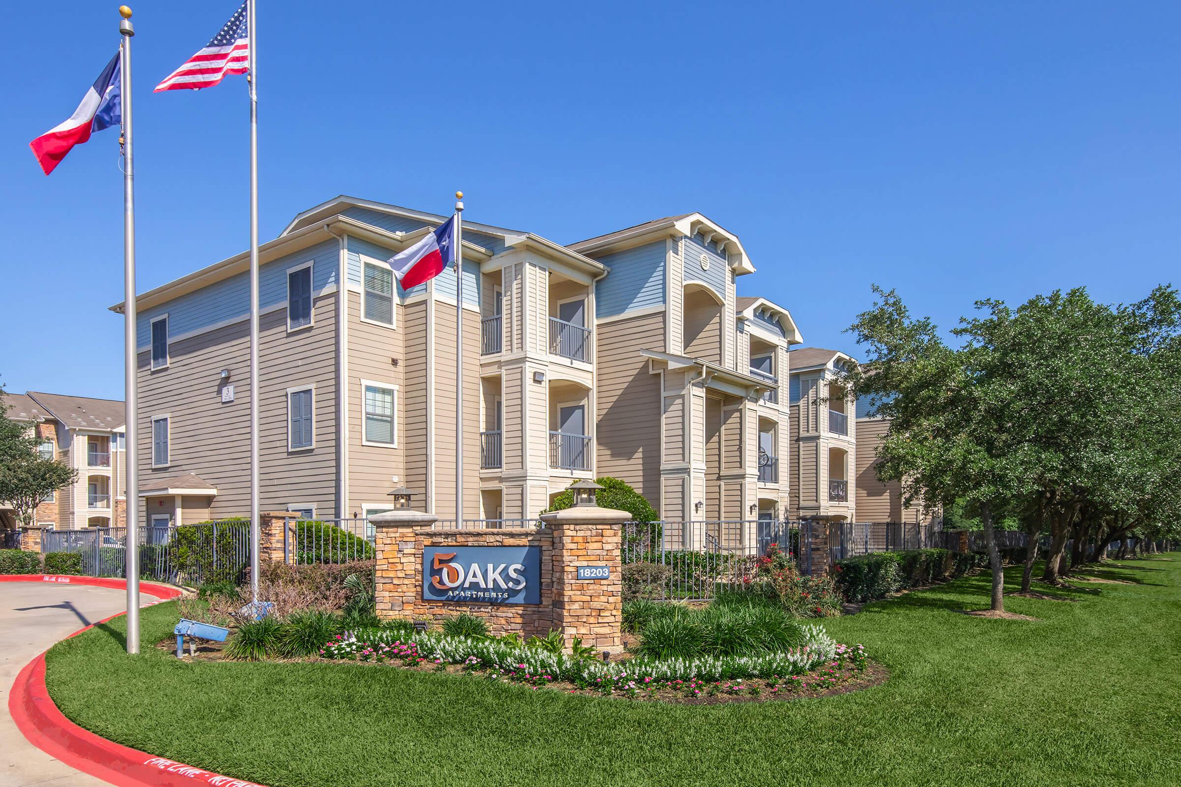 Exterior view of 5 Oaks Apartments featuring a well-maintained landscape with colorful flower beds, a sign at the entrance, and flags of the United States and Texas flying in front of the building. The building has multiple levels with balconies, surrounded by green grass and trees.