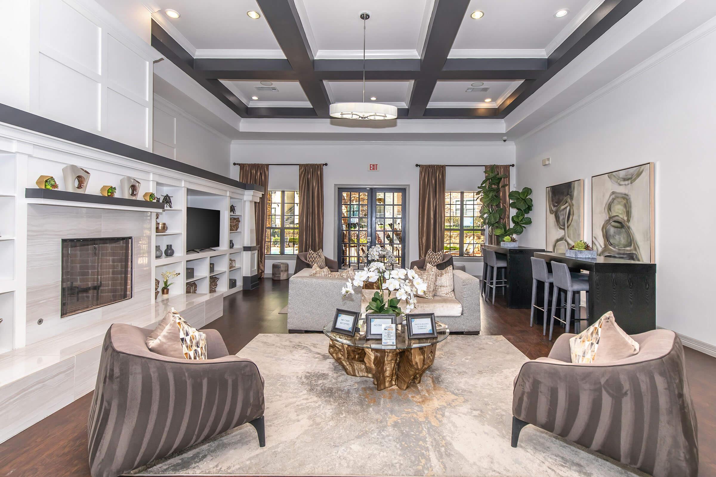 A spacious living area featuring elegant seating with plush chairs and a large area rug. The room is well-lit with a coffered ceiling and modern lighting fixtures. There is a decorative fireplace, a bookshelf, and large windows allowing natural light, complemented by a stylish dining area in the background. Plants add a touch of greenery.