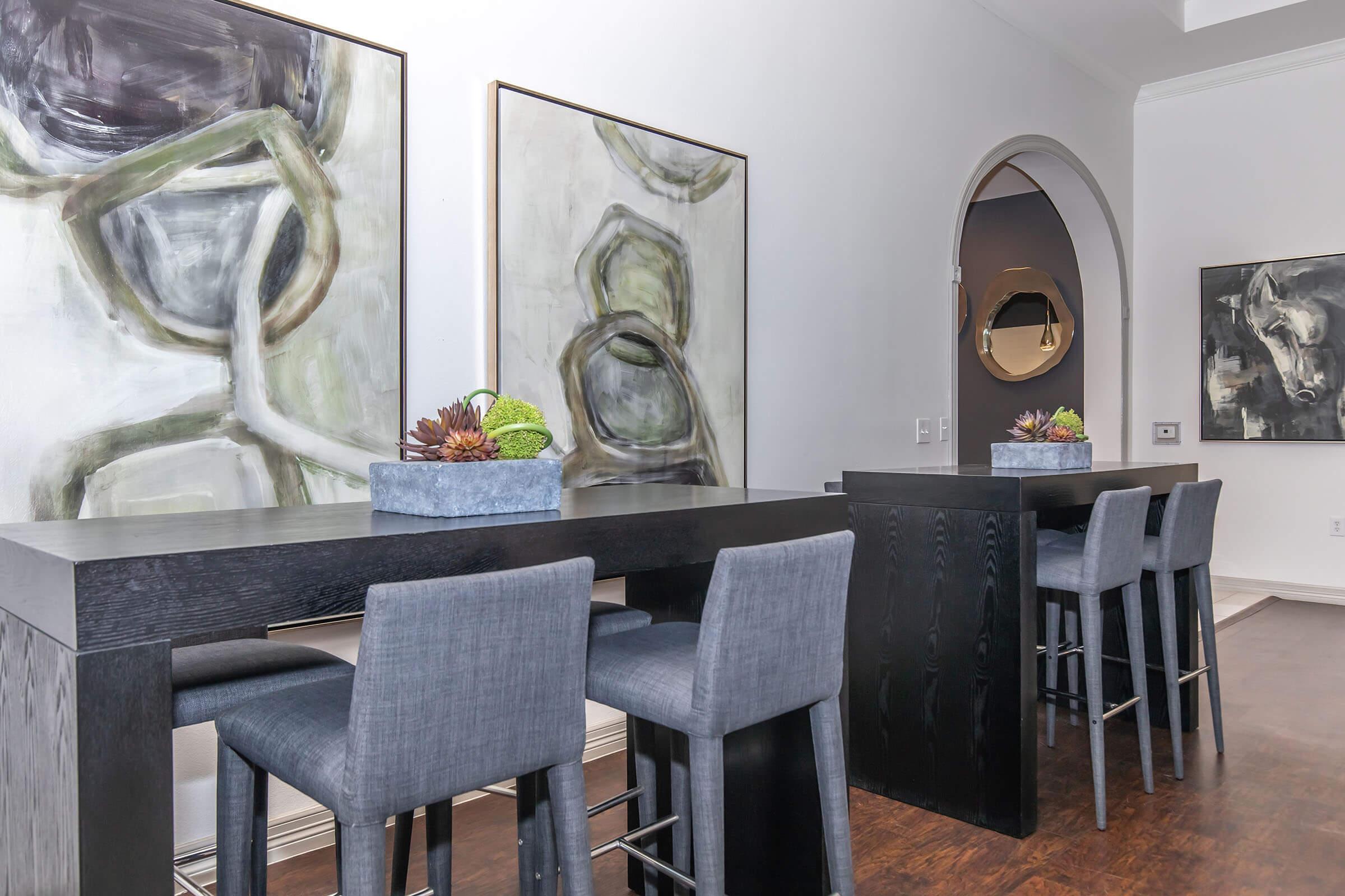 Modern interior featuring a stylish dining area with a long black table and grey upholstered chairs. Two abstract paintings with muted colors hang on the wall, and a decorative planter with succulents sits on the table. The space has a clean and contemporary aesthetic, illuminated by natural light.