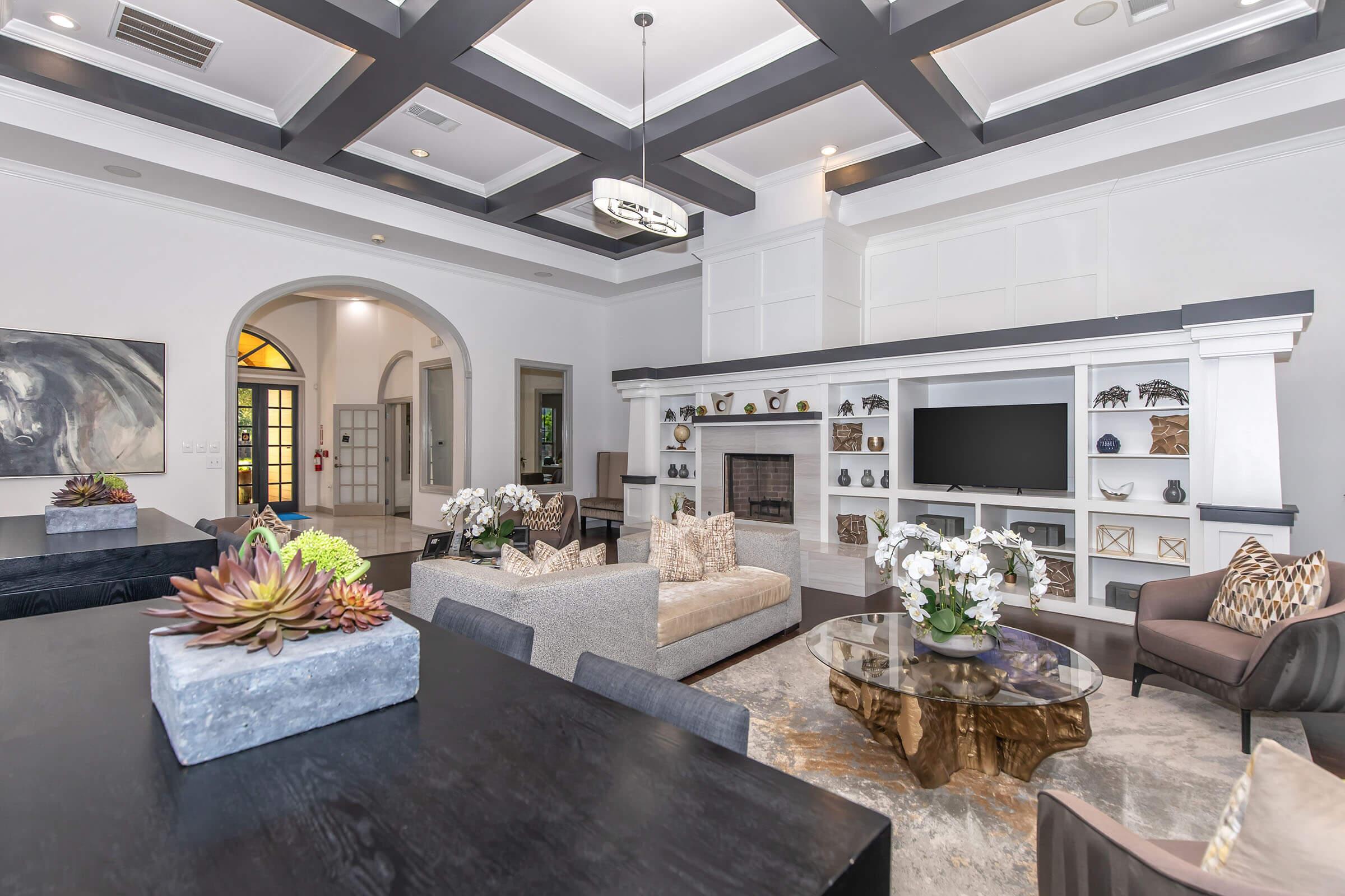 A spacious, elegantly designed living room featuring a cozy seating area with a sectional sofa and armchairs, a large coffee table, and decorative plants. The room has high ceilings with exposed beams, a modern fireplace, built-in shelves with decor, and a large window allowing natural light.