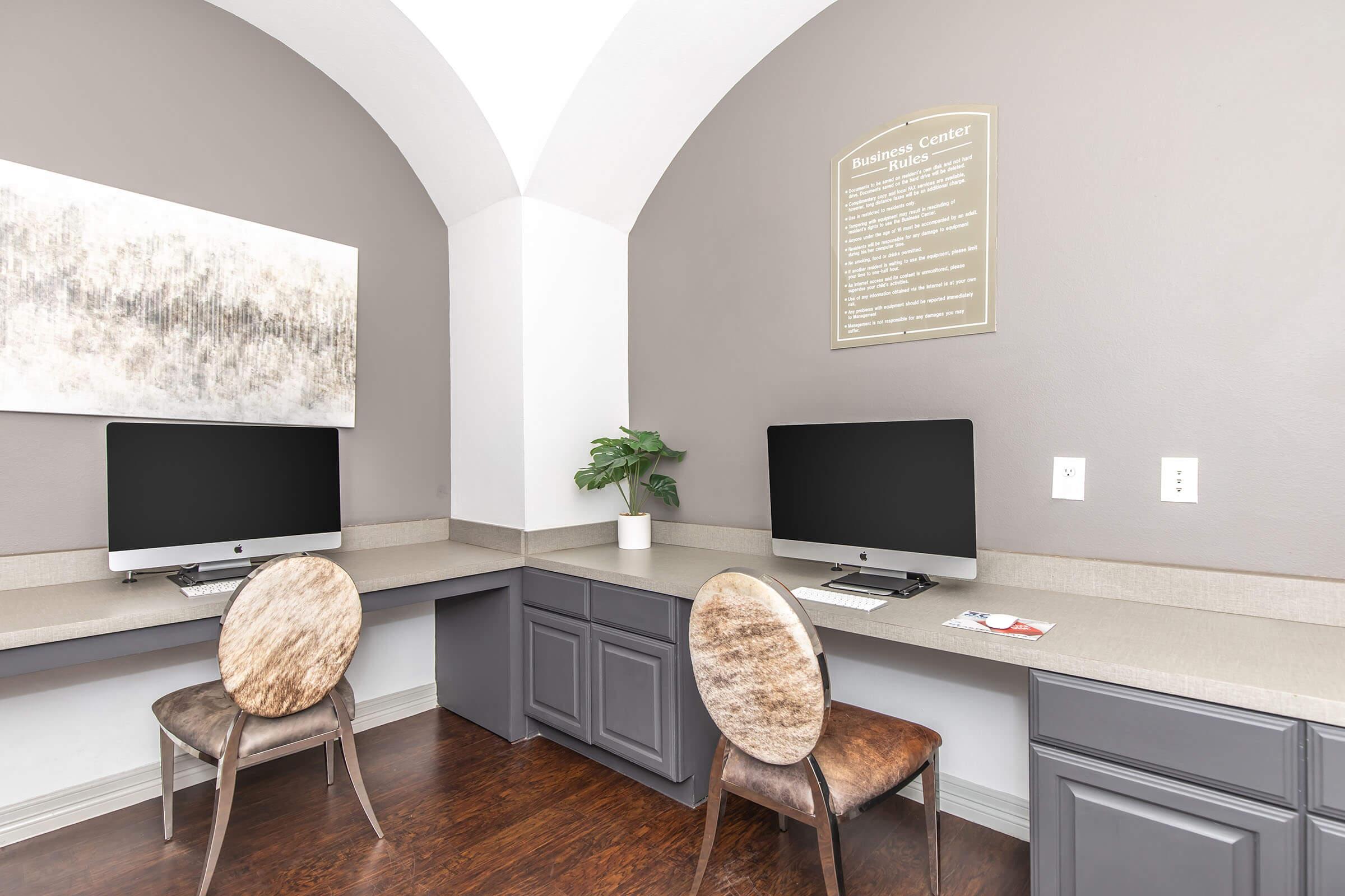 A modern workspace featuring two desktop computers on a gray countertop, accompanied by two stylish, round chairs. A potted plant adds a touch of greenery, while a large, abstract artwork hangs on the wall. The room is softly lit with an arched ceiling and neutral-colored walls. A sign is mounted above the workspace.
