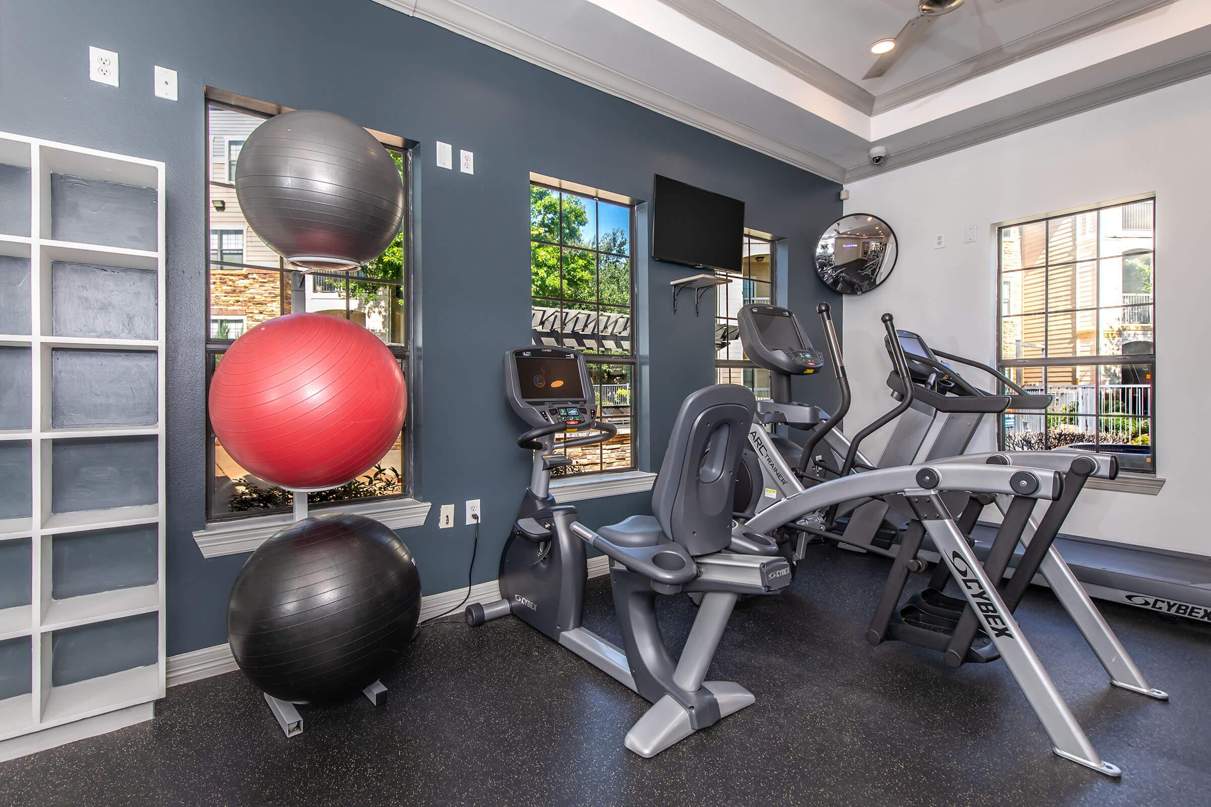 A modern gym interior featuring exercise equipment, including elliptical machines and stationary bikes. Colorful exercise balls are arranged against a wall. Large windows let in natural light, enhancing the welcoming atmosphere. A television and mirrors are also present, contributing to the fitness space's design.