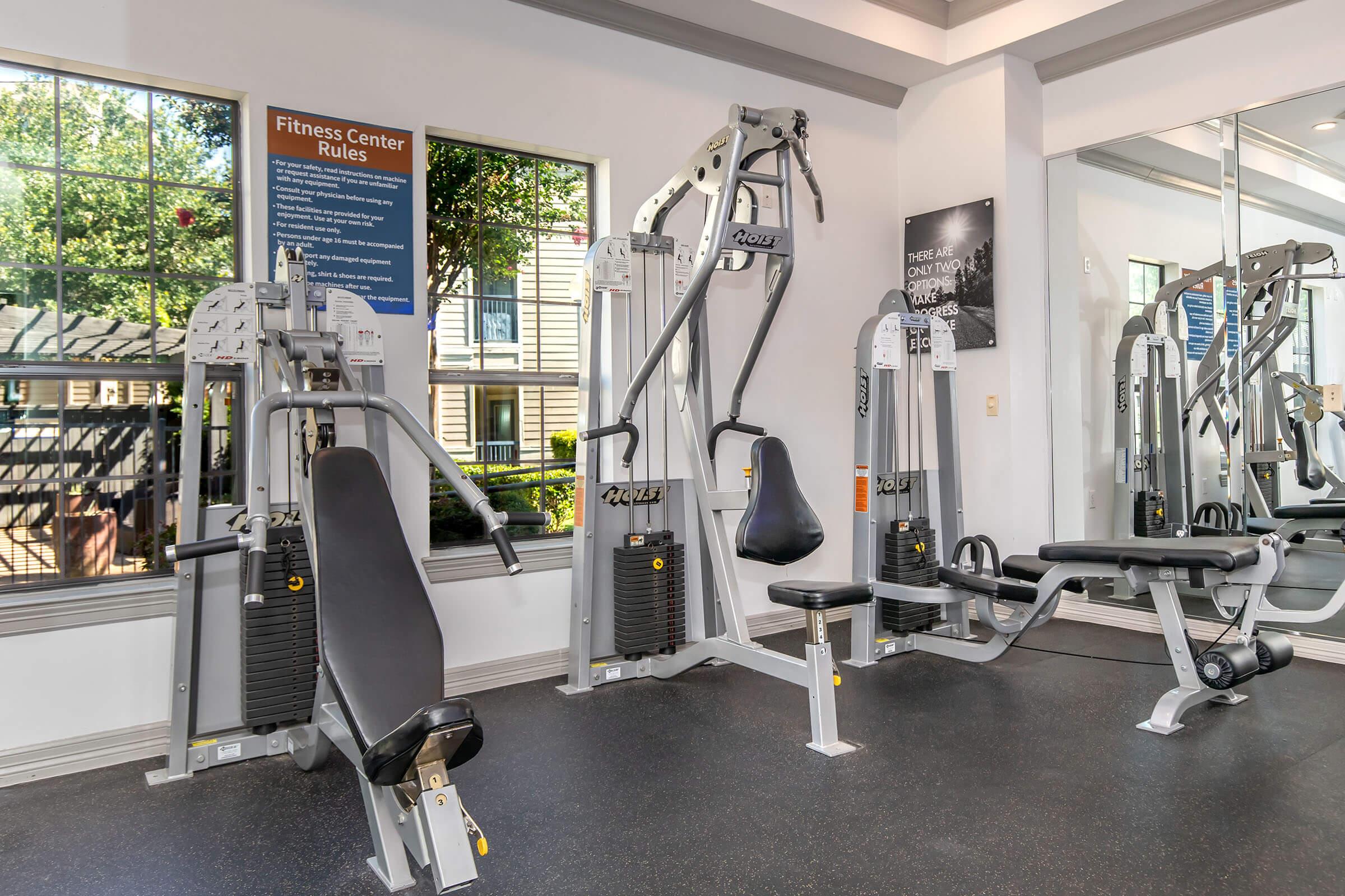 A well-equipped fitness center featuring various exercise machines, including a multi-station weight machine and a single-seat machine. Natural light comes through large windows, showcasing the outdoor environment. Fitness center rules are displayed on the wall.