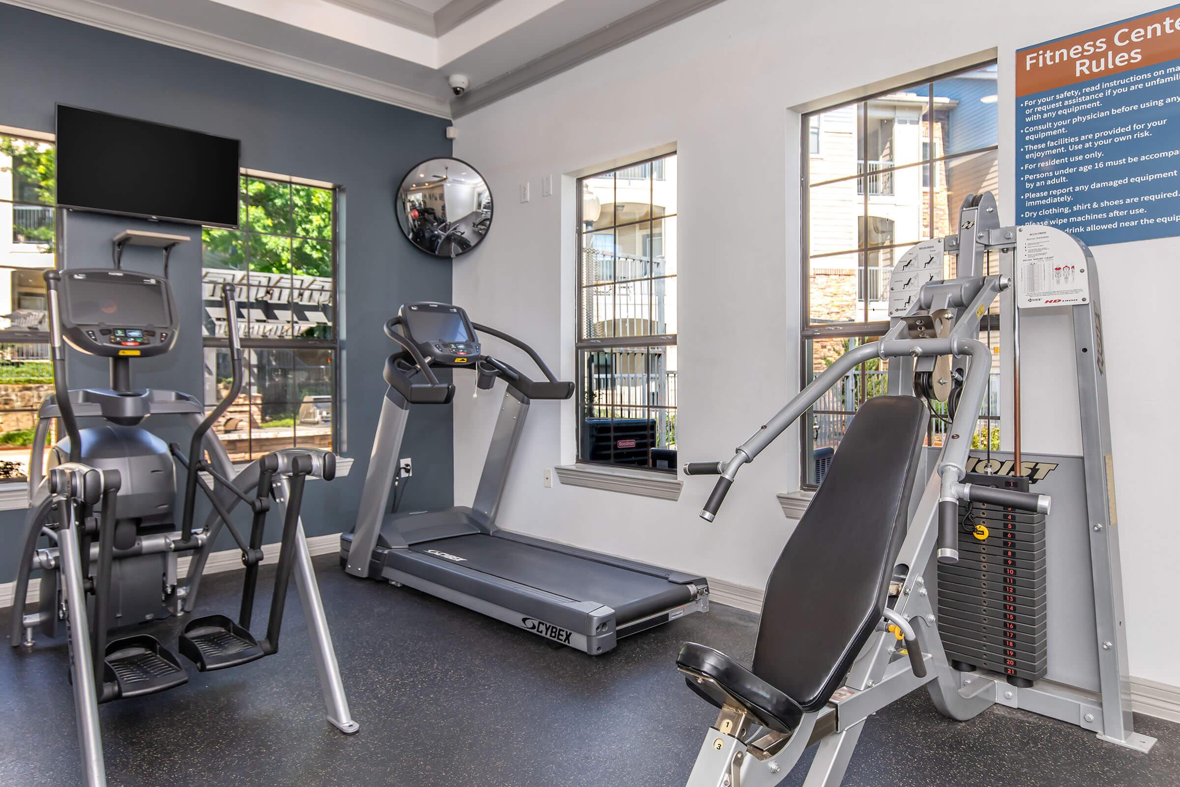 A well-lit fitness center featuring a treadmill, elliptical machine, and weight training equipment. Large windows provide natural light, and a wall sign outlines fitness center rules. A mirror is also visible, enhancing the sense of space.