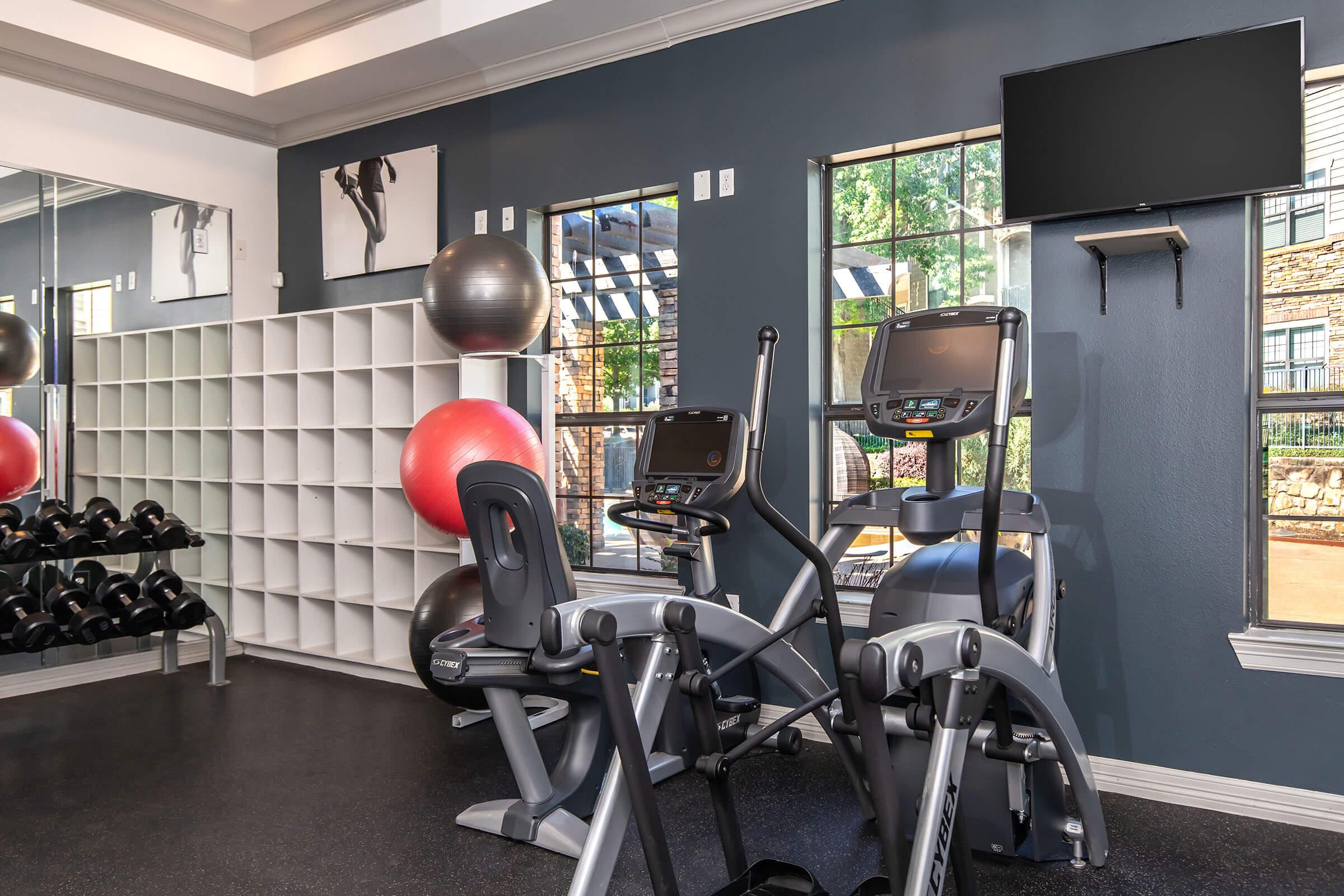 A modern gym interior featuring two exercise machines (a treadmill and an elliptical) positioned near large windows. There's a wall-mounted television and a rack of dumbbells. Red exercise balls are placed on the floor, enhancing the workout environment with good natural light streaming in.
