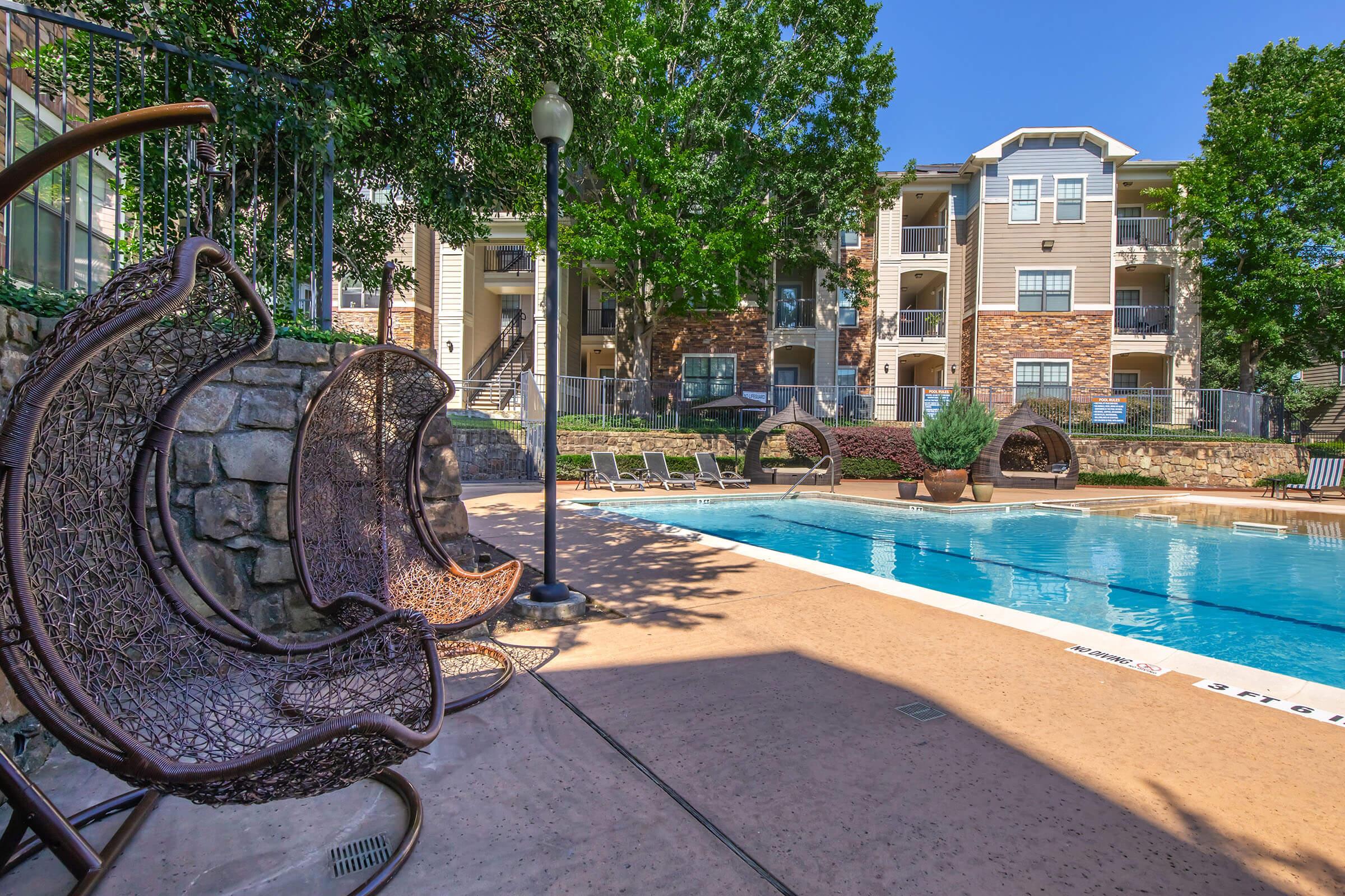 A view of an outdoor swimming pool surrounded by lounge chairs and decorative greenery. In the foreground, unique wicker chairs are positioned near the pool area, with an apartment building visible in the background. The sky is clear and blue, indicating a sunny day.