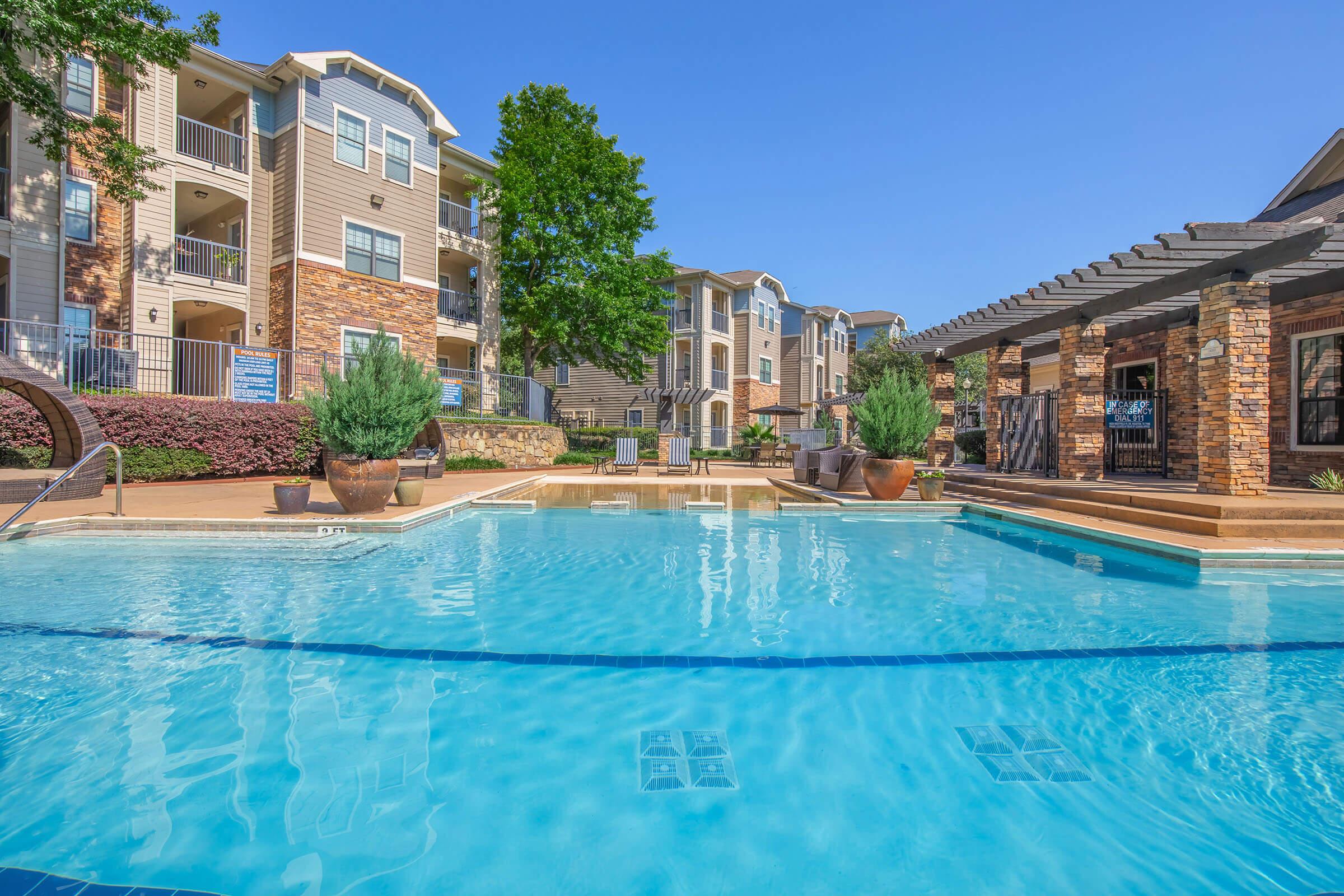 A clear blue swimming pool surrounded by beautifully landscaped greenery and modern apartment buildings. There are lounge chairs around the pool, and a pergola in the background, creating a serene and inviting atmosphere. The sky is bright and sunny, enhancing the appeal of the outdoor space.