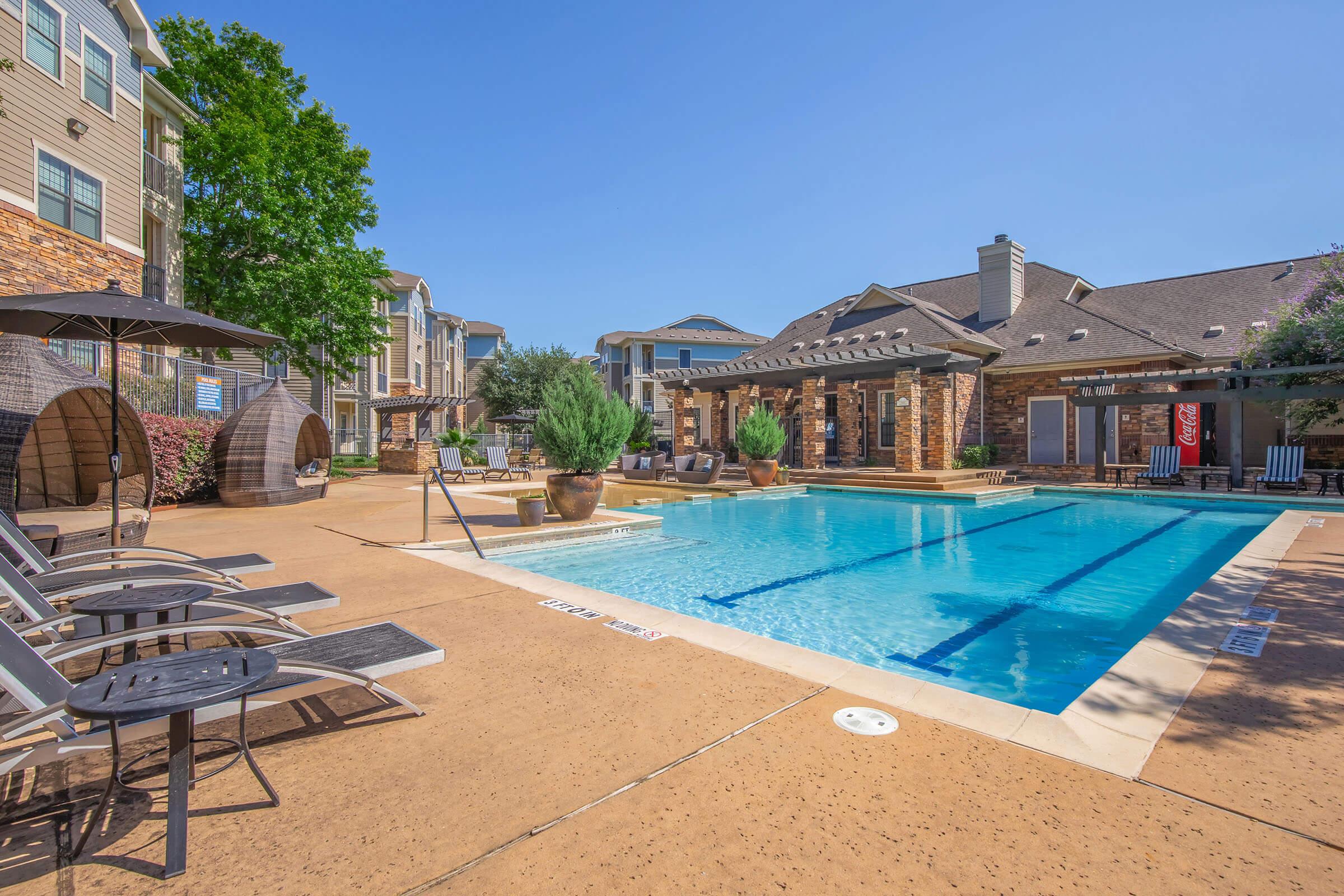 A sunny outdoor pool area with lounge chairs, a few potted plants, and shaded seating. Surrounding buildings have a modern design with brick and stone accents. Clear blue sky overhead and well-maintained landscaping create a relaxing atmosphere.