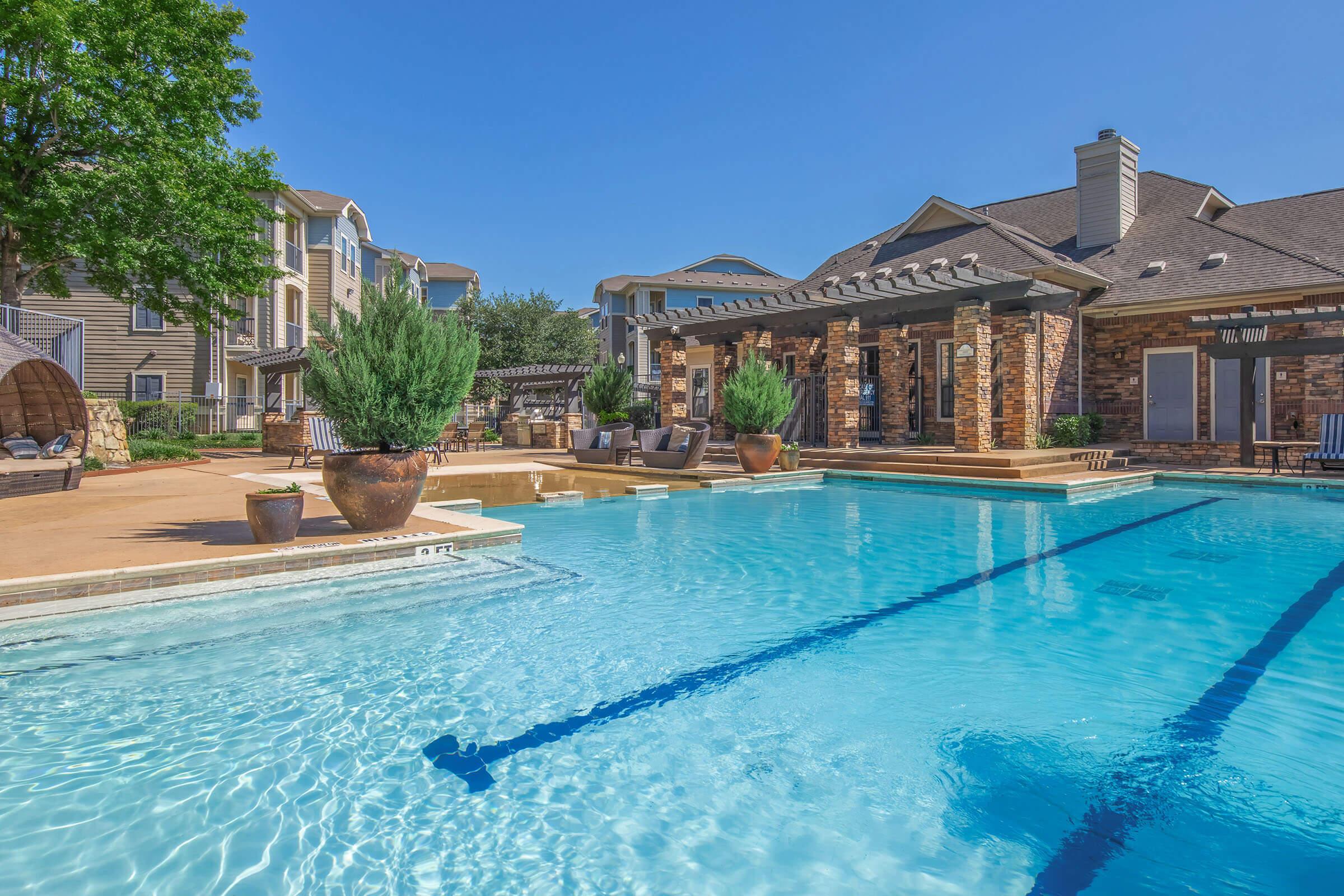 A bright swimming pool surrounded by stone patios and lush greenery. The pool features lanes for swimming, with decorative plants in large pots. In the background, there's a stylish building with a pergola, providing a relaxing atmosphere under a clear blue sky.