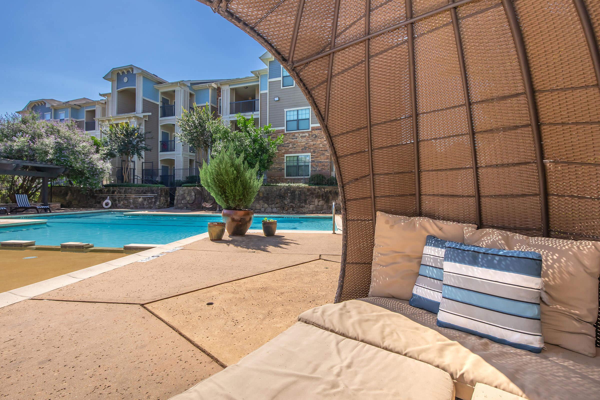 Cozy seating area with plush cushions under a woven canopy near a swimming pool. Lush greenery and potted plants are visible in the background, alongside modern apartment buildings. Clear blue sky overhead adds to the inviting atmosphere of this outdoor relaxation space.