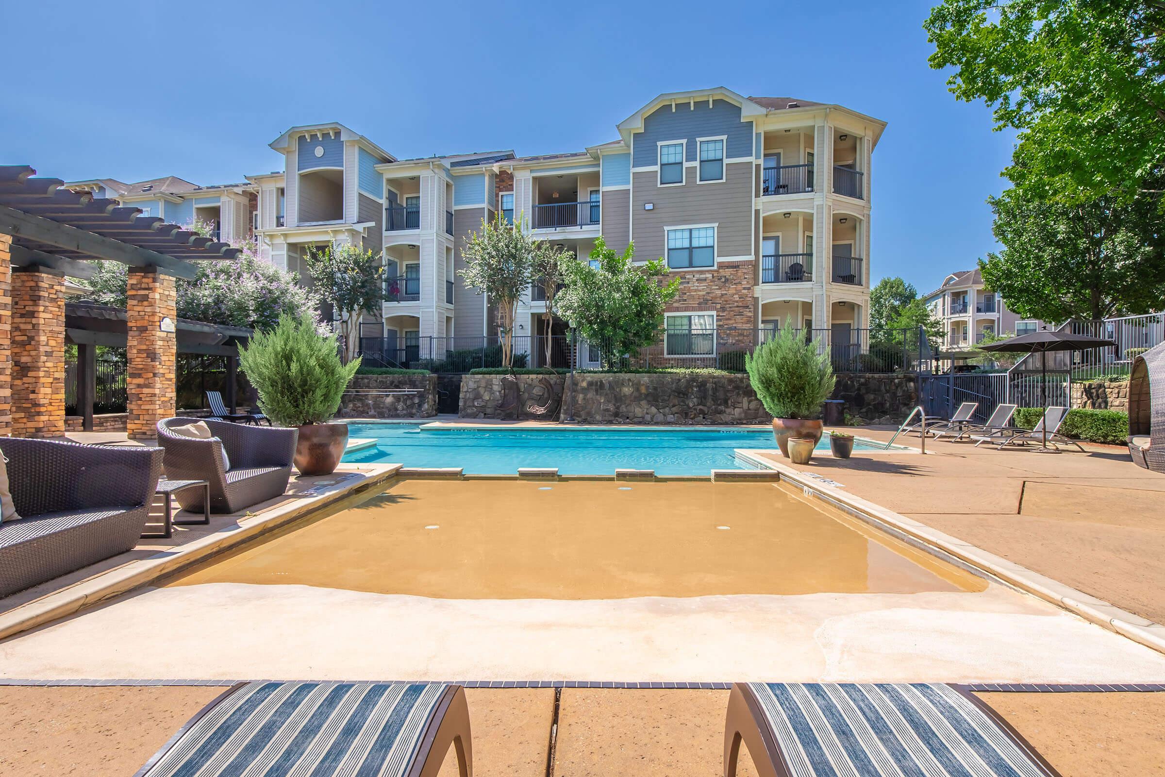 A sunny poolside view featuring a tranquil swimming pool surrounded by lounge chairs and potted plants. In the background, there are modern apartment buildings with balconies, and a clear blue sky adds to the inviting atmosphere of the outdoor space.