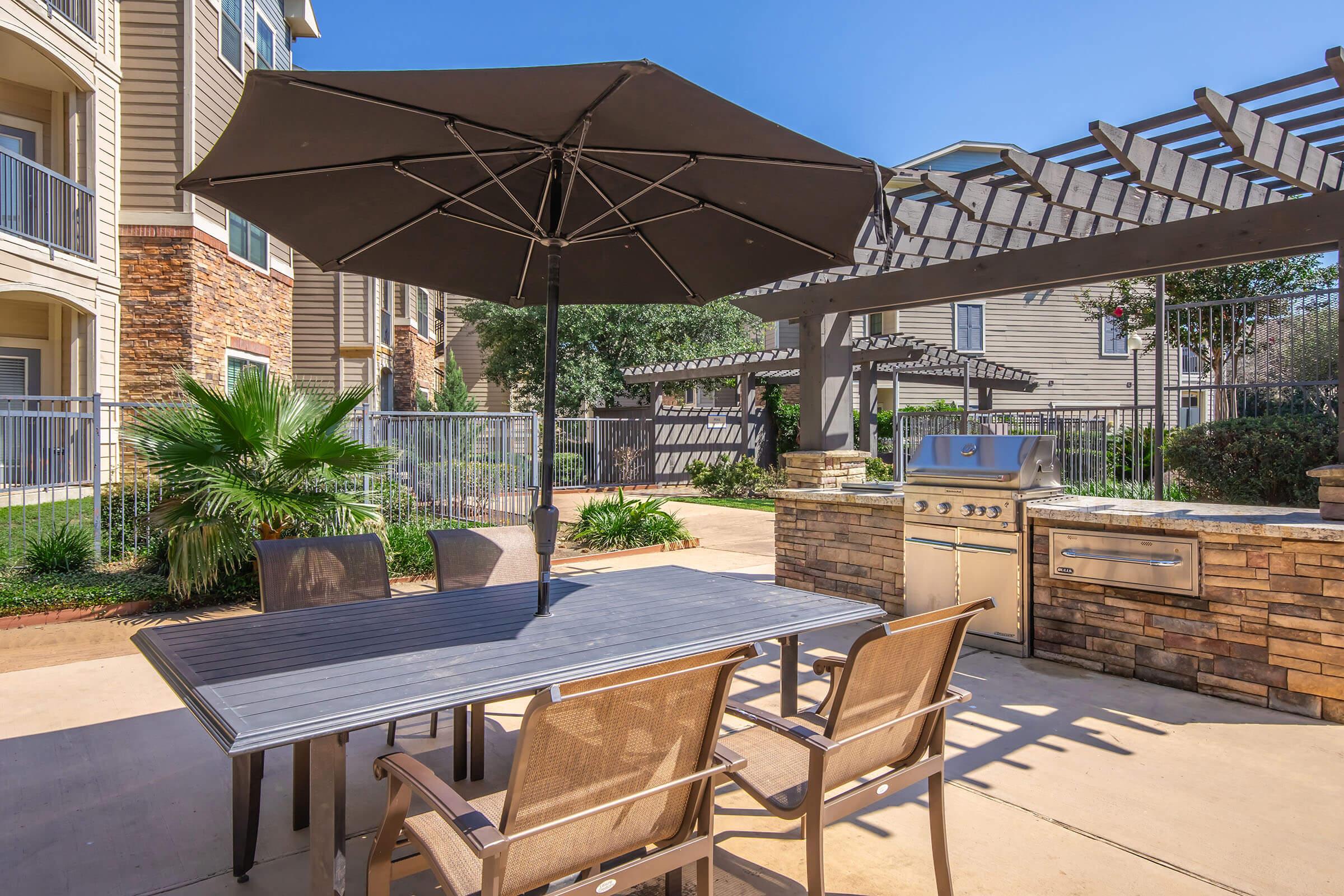 A modern outdoor dining area featuring a large umbrella over a rectangular table surrounded by four chairs. In the background, there is a stone barbecue grill, a pergola, and lush greenery, creating a welcoming atmosphere for social gatherings in a residential complex.