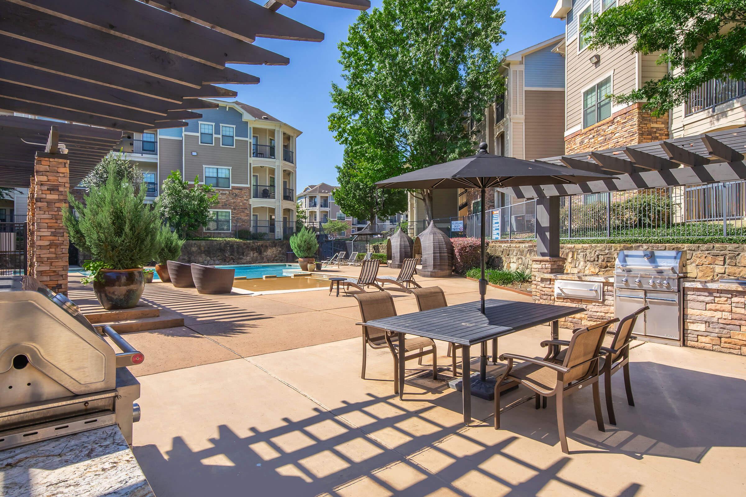 A modern outdoor pool area featuring lounge chairs, a dining table under an umbrella, a grill, and decorative plants. Surrounding the pool are apartment buildings, and the space is sunny with clear blue skies. The atmosphere is inviting for leisure and relaxation.