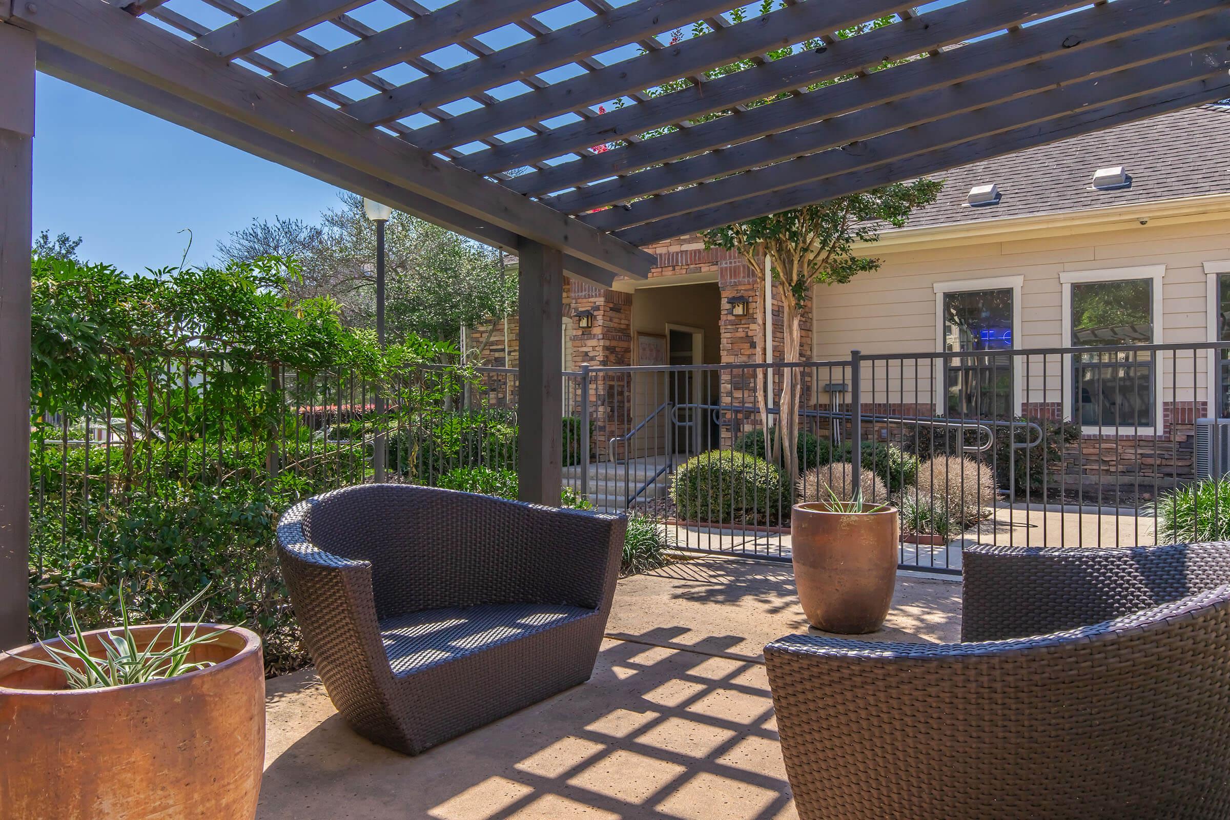 A shaded outdoor seating area featuring two woven lounge chairs, surrounded by green shrubs and potted plants. A stone building is visible in the background, along with a wrought-iron fence. The scene is bright and inviting, suitable for relaxation.