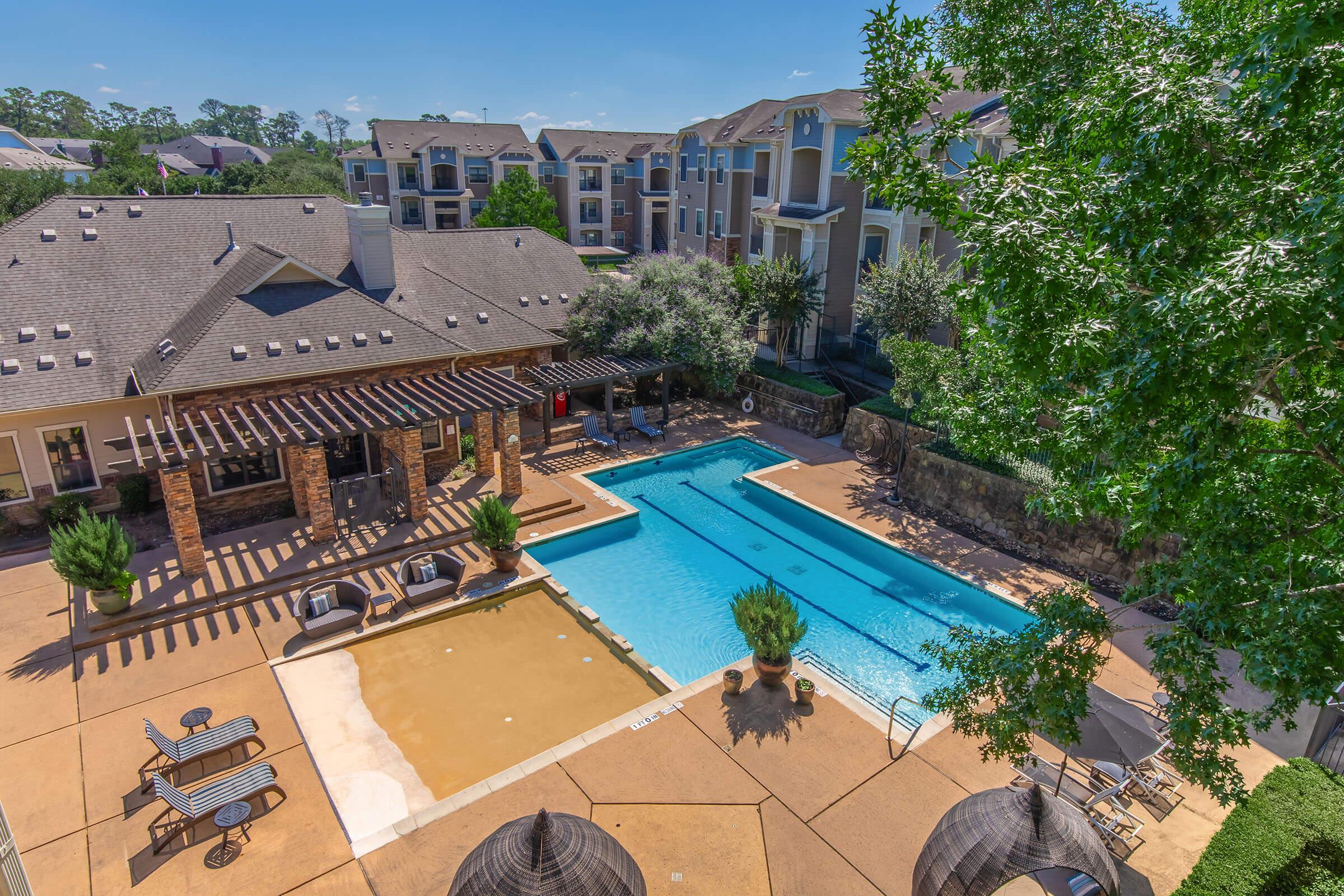 Aerial view of a landscaped pool area featuring a large swimming pool, lounge chairs, and shaded seating under a pergola. Surrounding the pool are lush trees and well-maintained greenery, with apartment buildings visible in the background against a clear blue sky.