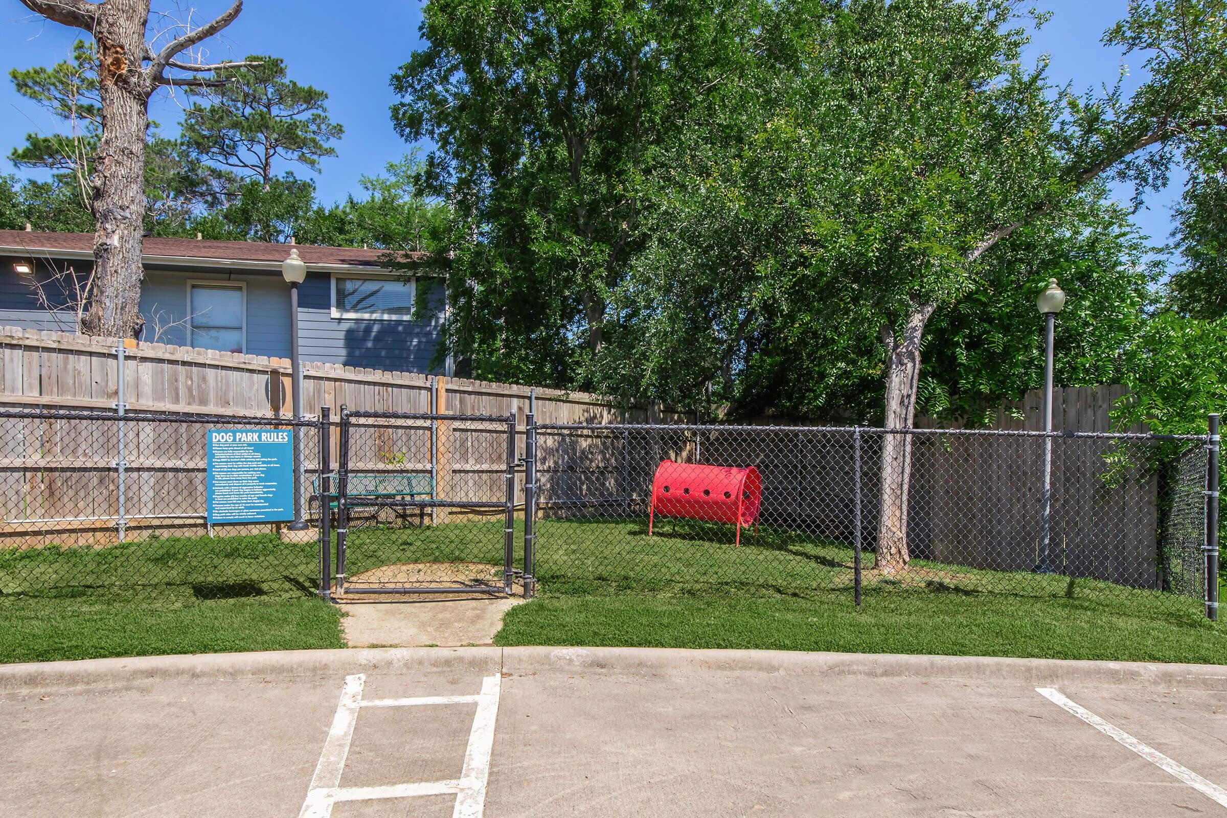 A fenced dog park with green grass, featuring a red dog play tunnel and a sign with rules. There are trees and a wooden fence in the background, and a parking area in the foreground. Bright blue skies are visible above the park.