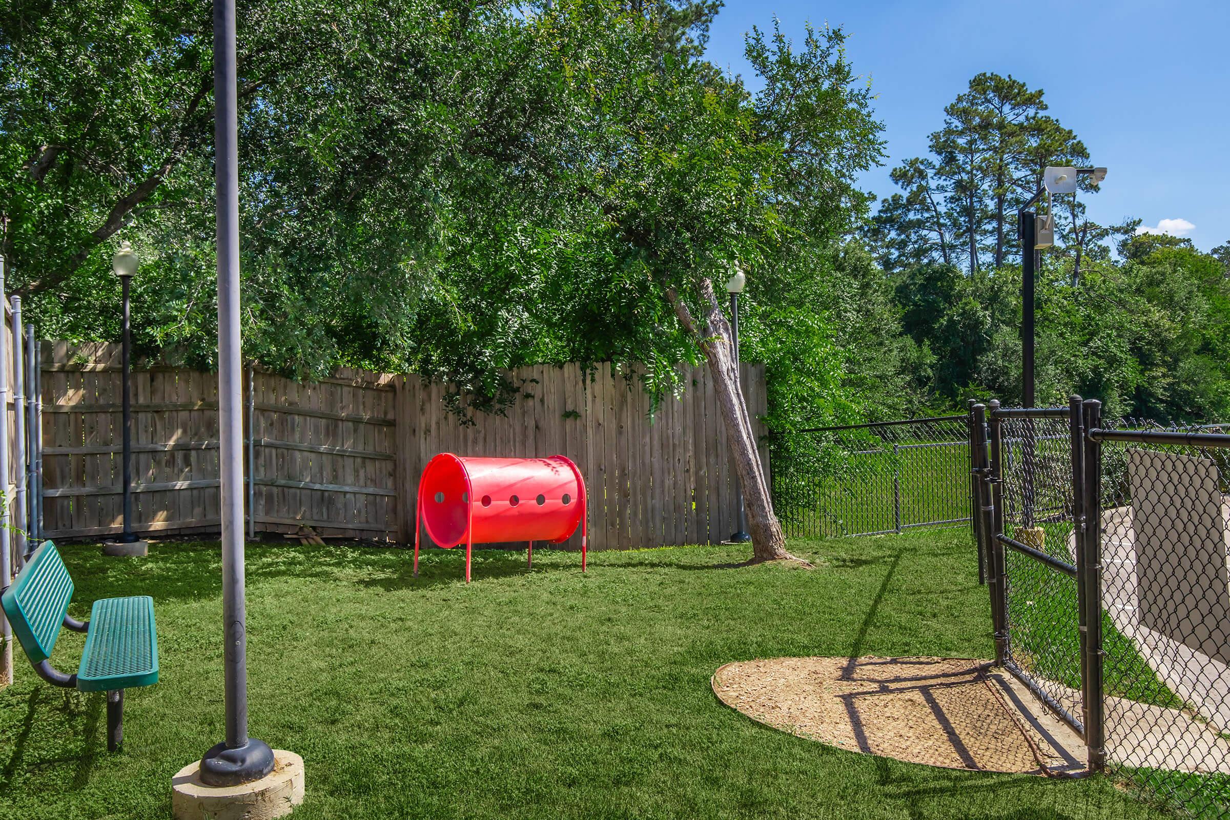 A bright outdoor area with a grassy surface, featuring two green benches and a red cylindrical play structure. A wooden fence and chain-link fence enclose the space, with trees and a clear blue sky in the background, creating a pleasant environment for recreation.