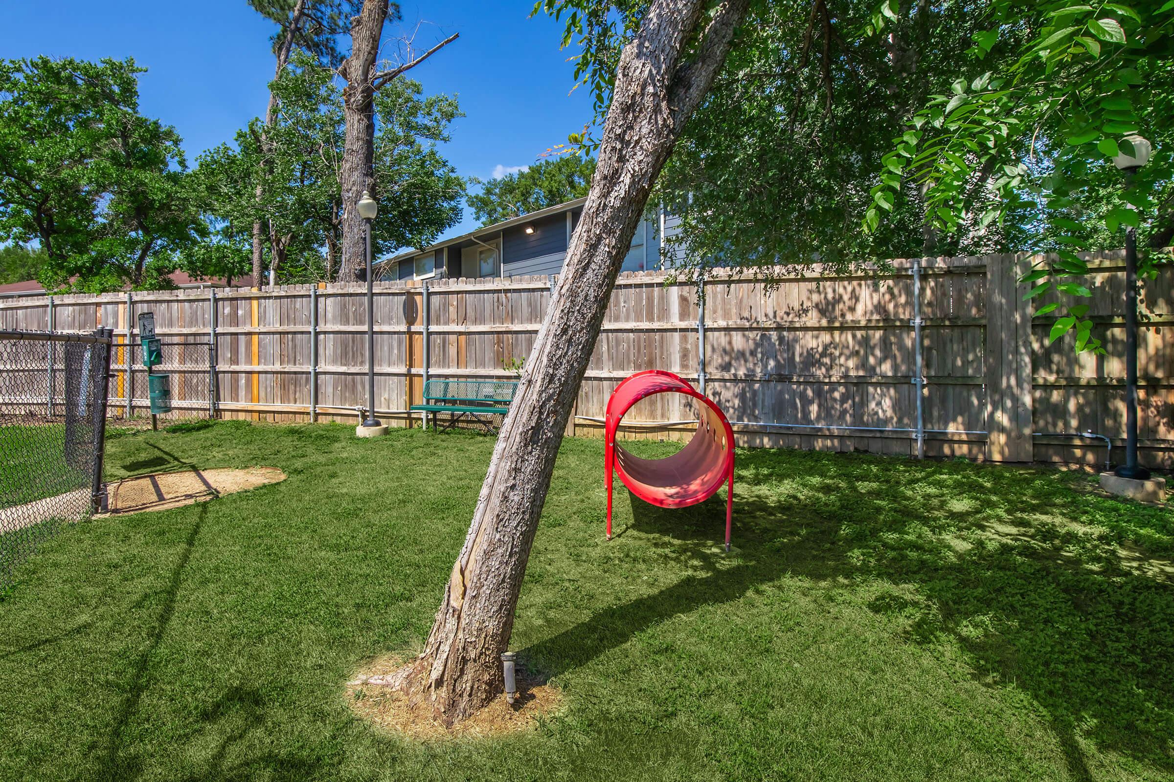 A sunny backyard featuring a well-maintained lawn, a large tree, and a bright red circular play tunnel. A wooden fence surrounds the area, and there's a bench in the background, offering a peaceful outdoor space. The sky is clear with few clouds.