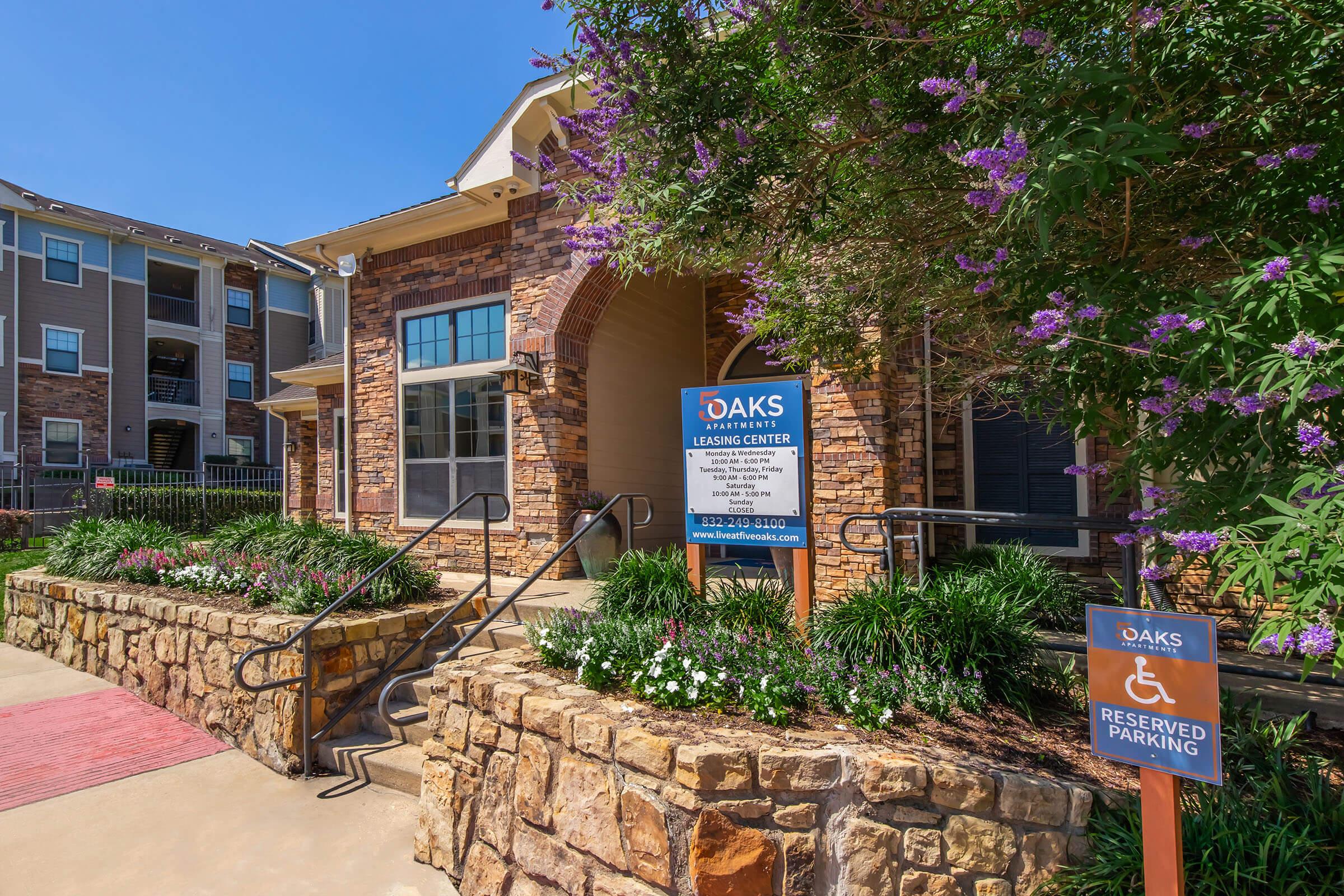 A modern building entrance with stone accents, surrounded by colorful flowers and landscaping. A sign for "Oaks Learning Center" is prominently displayed. In front, a reserved parking sign is visible, indicating designated parking. The atmosphere is welcoming and well-maintained.