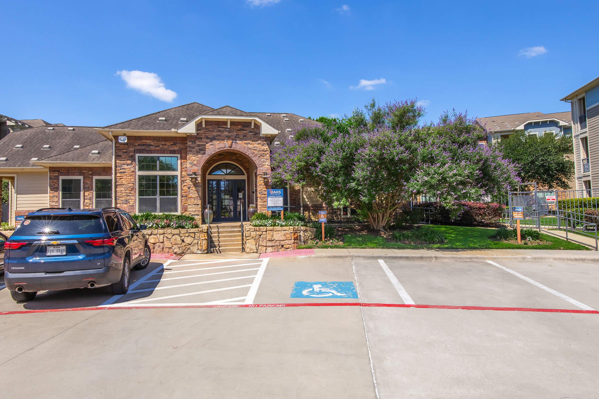 A stone and brick building with large windows and a welcoming entrance, surrounded by green trees and landscaping. In the foreground, a parking area features a designated handicap spot and a vehicle parked nearby. The sky is clear and blue, with a few clouds.