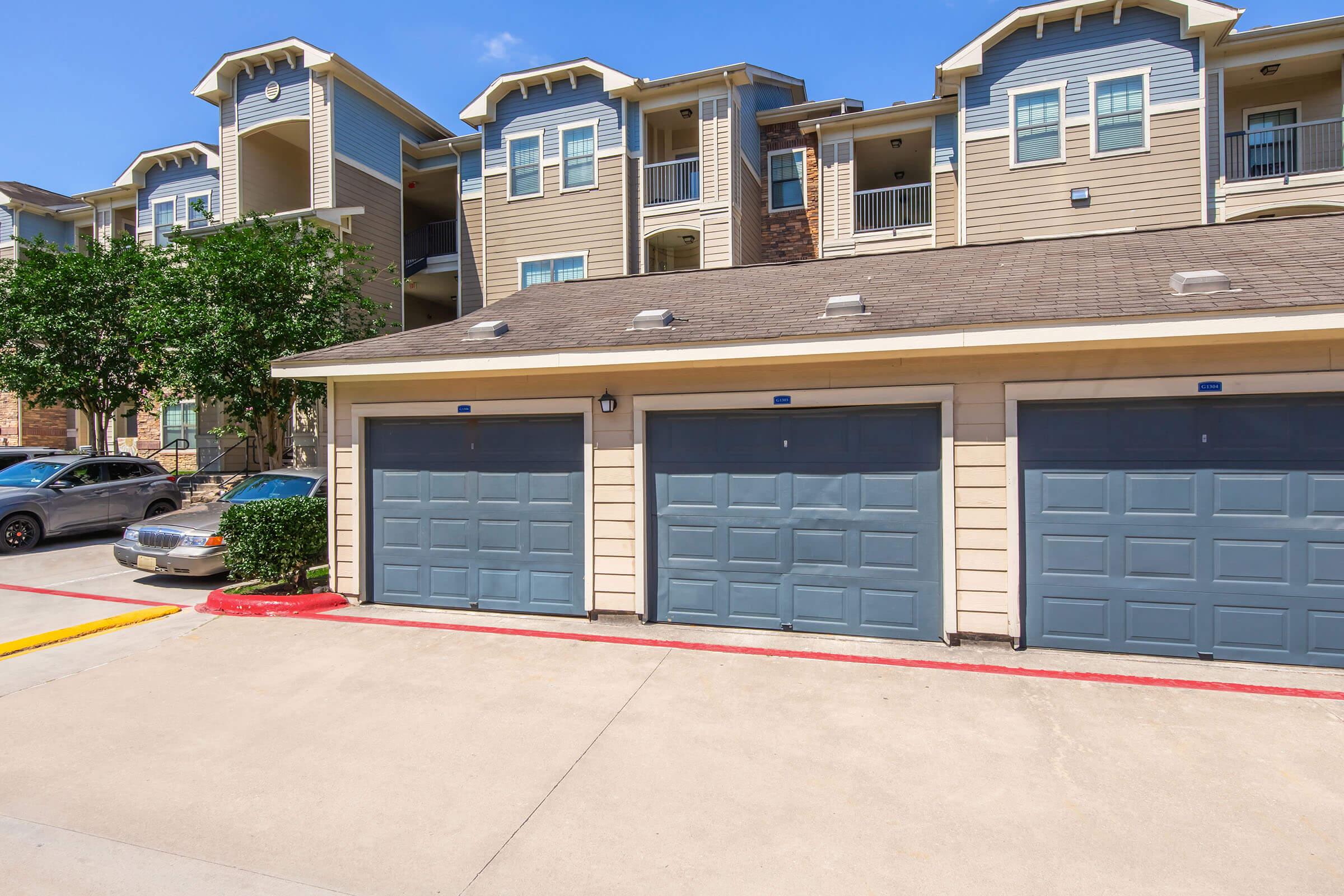 Two closed garages with gray doors are visible in the foreground, along with a parked car on the left. In the background, there are multi-story residential apartment buildings with a mix of beige and brown siding, and greenery is present beside the garages. The sky is clear and blue.