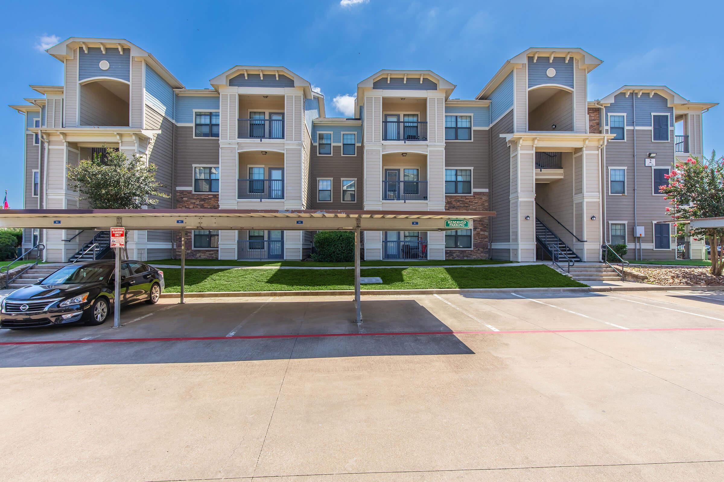 A modern apartment building featuring multiple floors with balconies and windows. The exterior is a combination of gray and beige siding, and there is a parking area with a car parked beneath a covered space. Green grass and shrubs are landscaped in front of the building. The sky is clear and blue.
