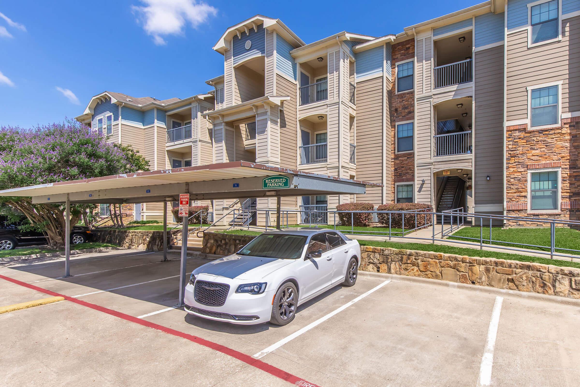 A white car parked under a covered parking space in front of a multi-story apartment building with a stone and wood exterior. The building has balconies and well-maintained landscaping with trees. The sky is clear and sunny, indicating a pleasant day.