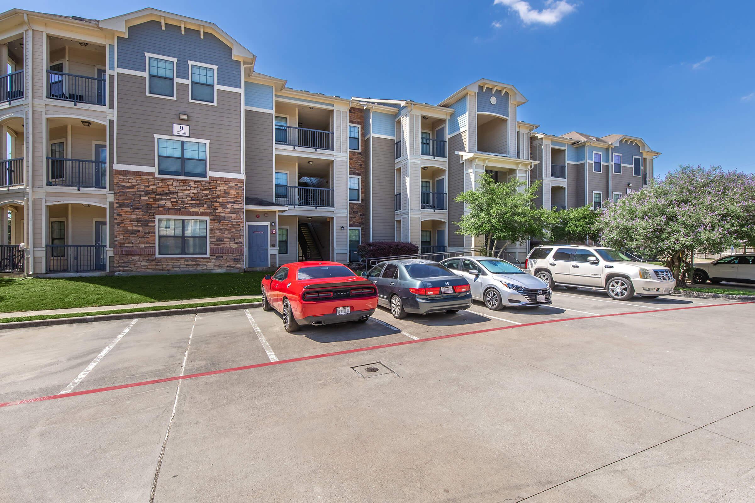 A multi-story apartment building with a mixture of stone and siding exteriors. In the foreground, there are several parked cars including a red sports car and a silver SUV. The building has balconies and green landscaping with trees, under a clear blue sky.