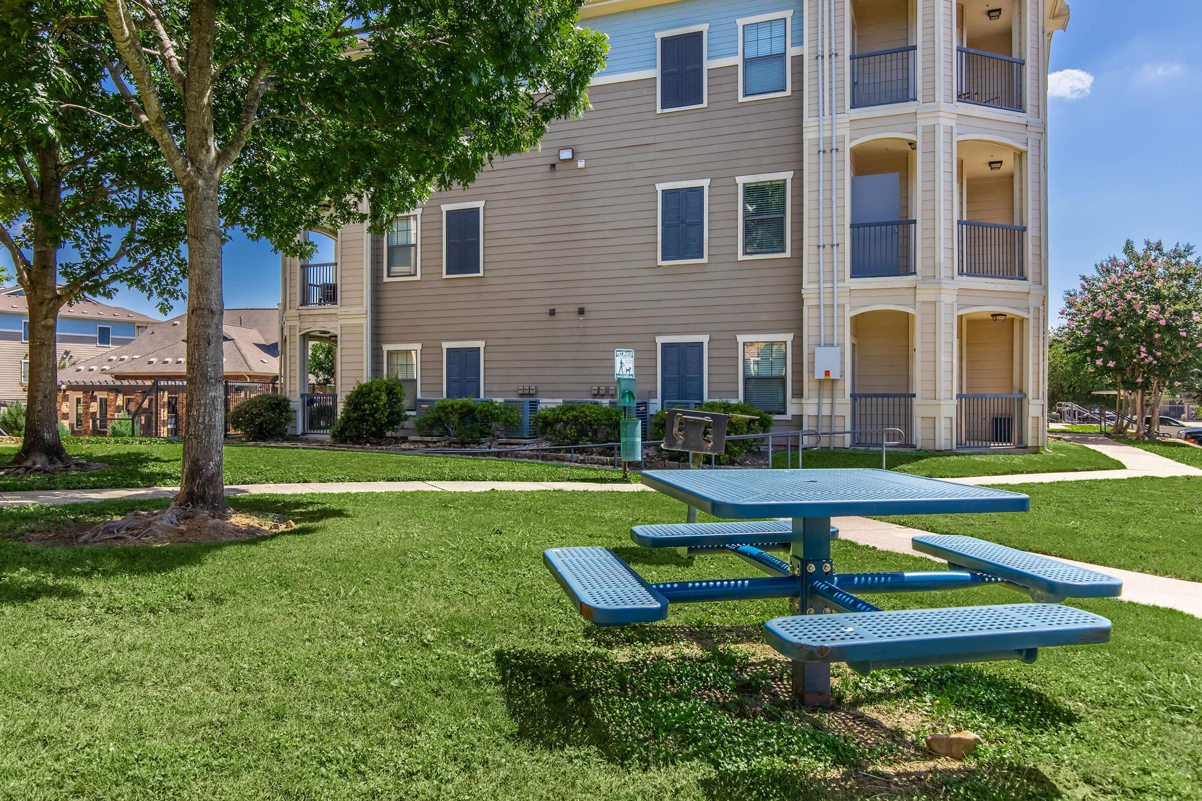 A picnic table with blue benches is situated on a grassy area in front of a modern multi-story apartment building. Trees provide shade, and there are pathways leading through the lawn. The sky is clear and sunny, creating a pleasant outdoor environment.