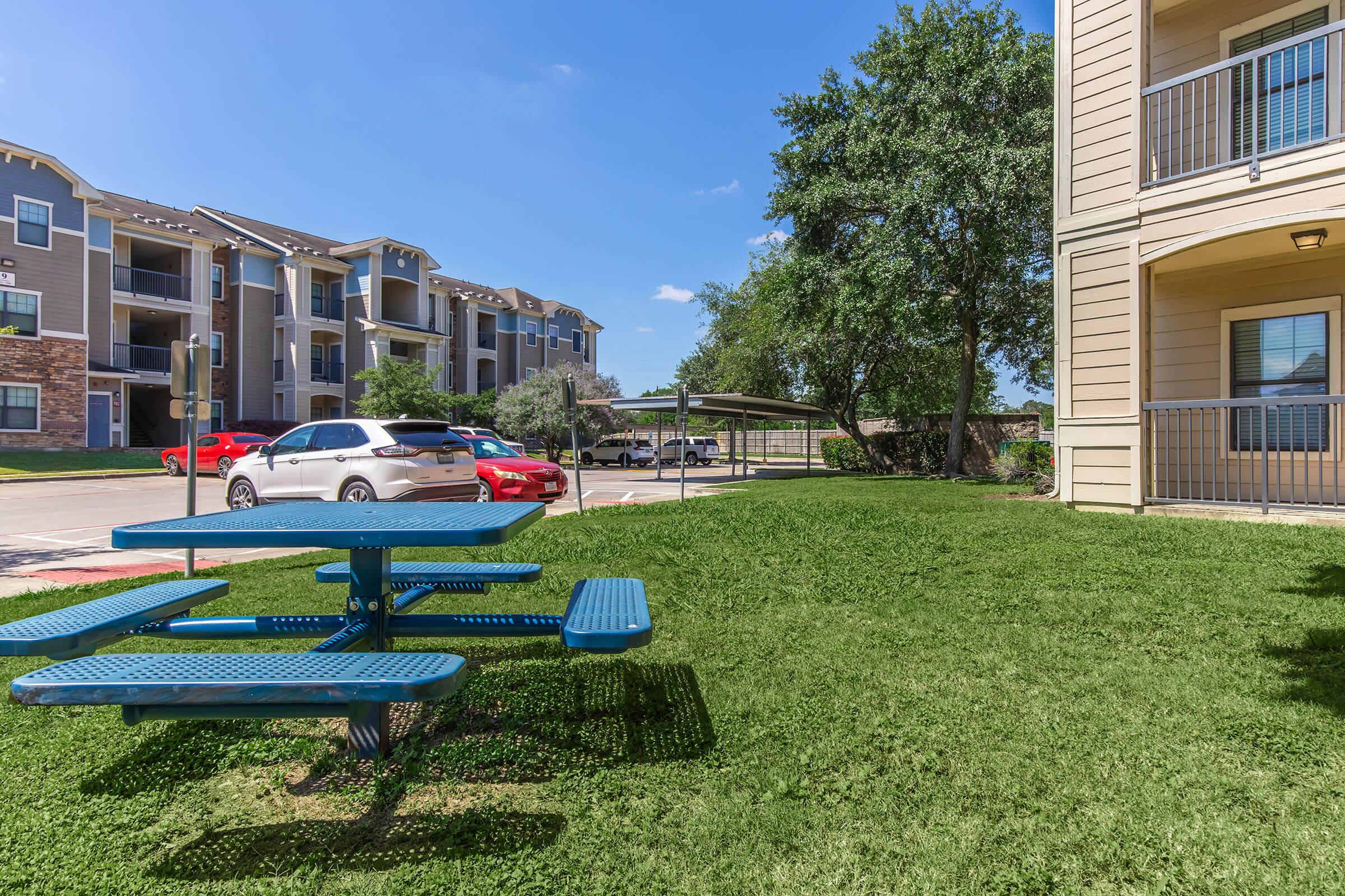 A blue picnic table sits on a grassy area in front of a multi-story apartment complex. Several parked cars are visible nearby, partially shaded by trees, with a clear blue sky overhead. The scene conveys a relaxed outdoor space adjacent to residential buildings.