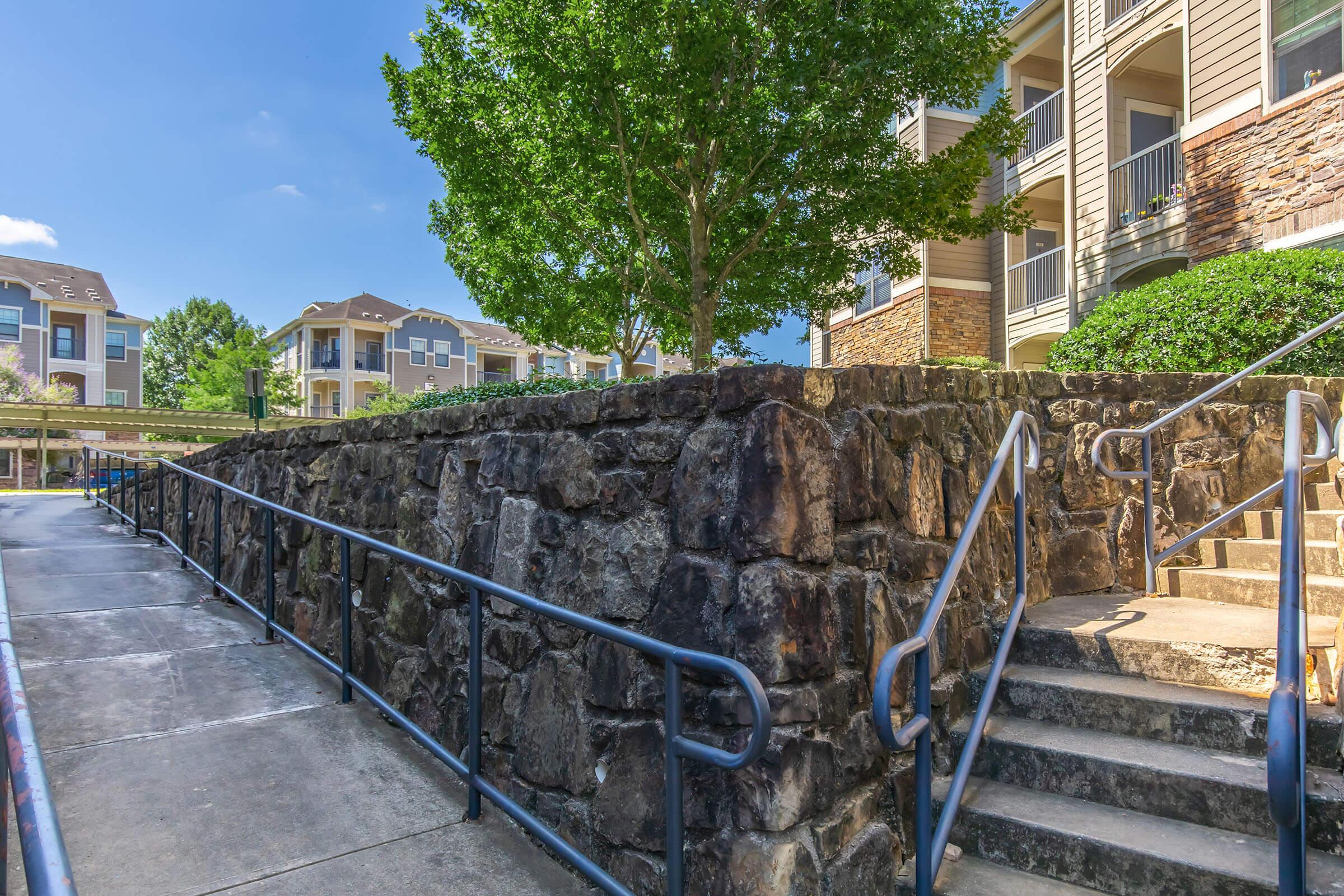 A pathway leading up to a stone wall with a tree on top, surrounded by apartment buildings. The scene features a clear blue sky and greenery, with an accessible ramp on one side and stairs on the other, creating a welcoming entryway to the residential area.