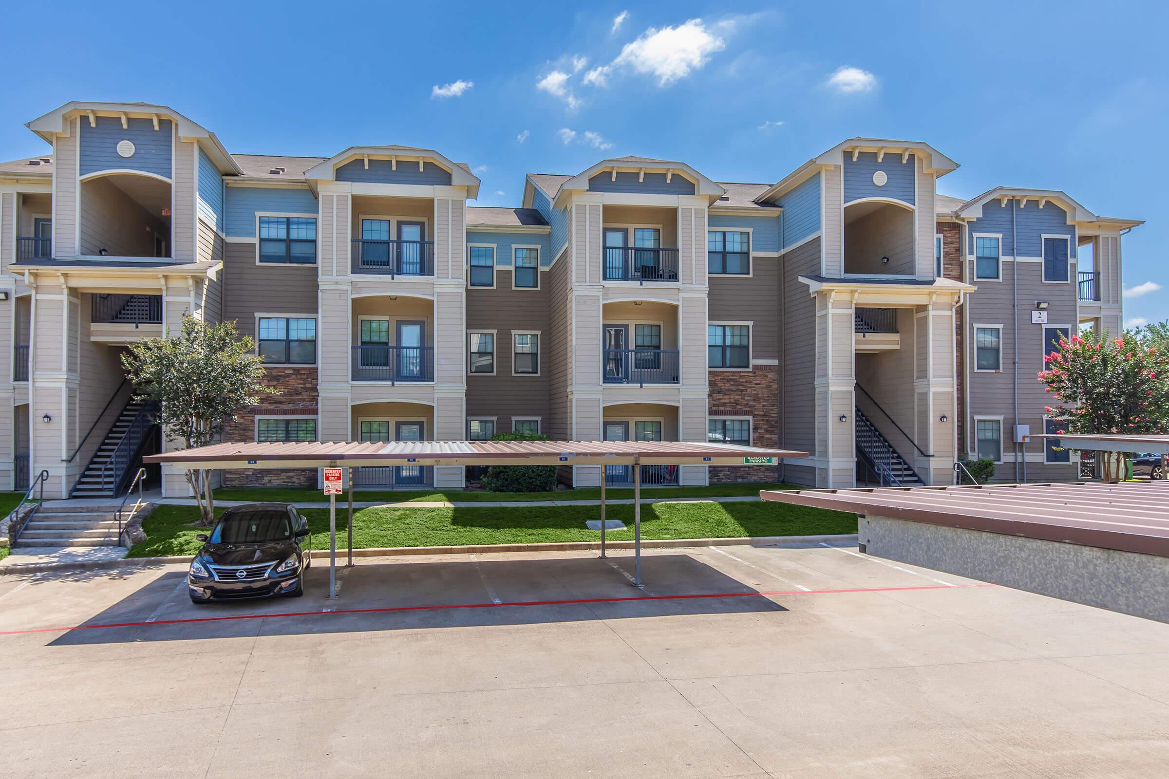 A modern multi-story apartment building with a mix of brick and siding. It features balconies on each unit, surrounded by green landscaping. A covered parking area is visible in the foreground, with a vehicle parked underneath. The sky is clear with a few clouds, indicating a sunny day.