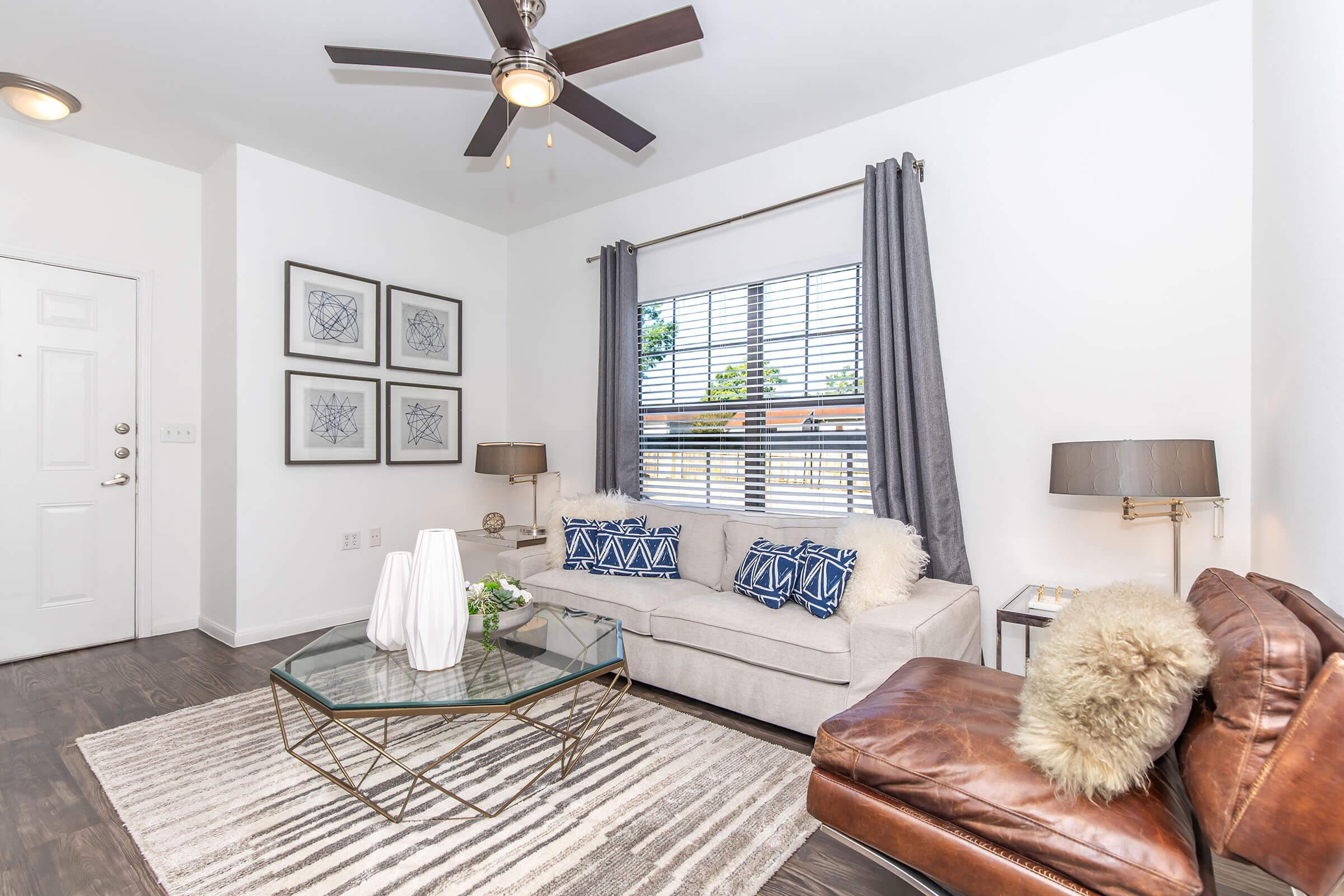 A cozy living room featuring a light gray sofa adorned with decorative blue pillows. A sleek glass coffee table sits on a patterned rug, and there’s a brown leather chair nearby. The room is brightened by a ceiling fan and natural light streaming through a window with gray curtains, framed by modern wall art.
