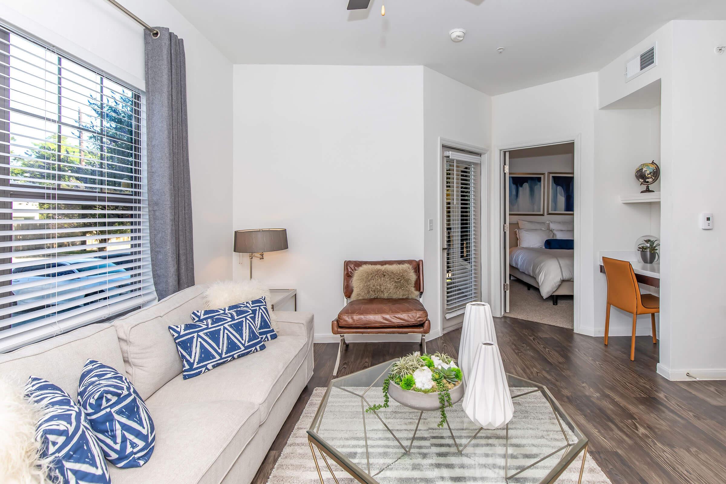 A cozy living room featuring a light beige sofa with patterned blue pillows, a stylish brown leather armchair, and a modern glass coffee table with a plant centerpiece. Natural light streams in through large windows with gray curtains, and a doorway leads to a bedroom area visible in the background.