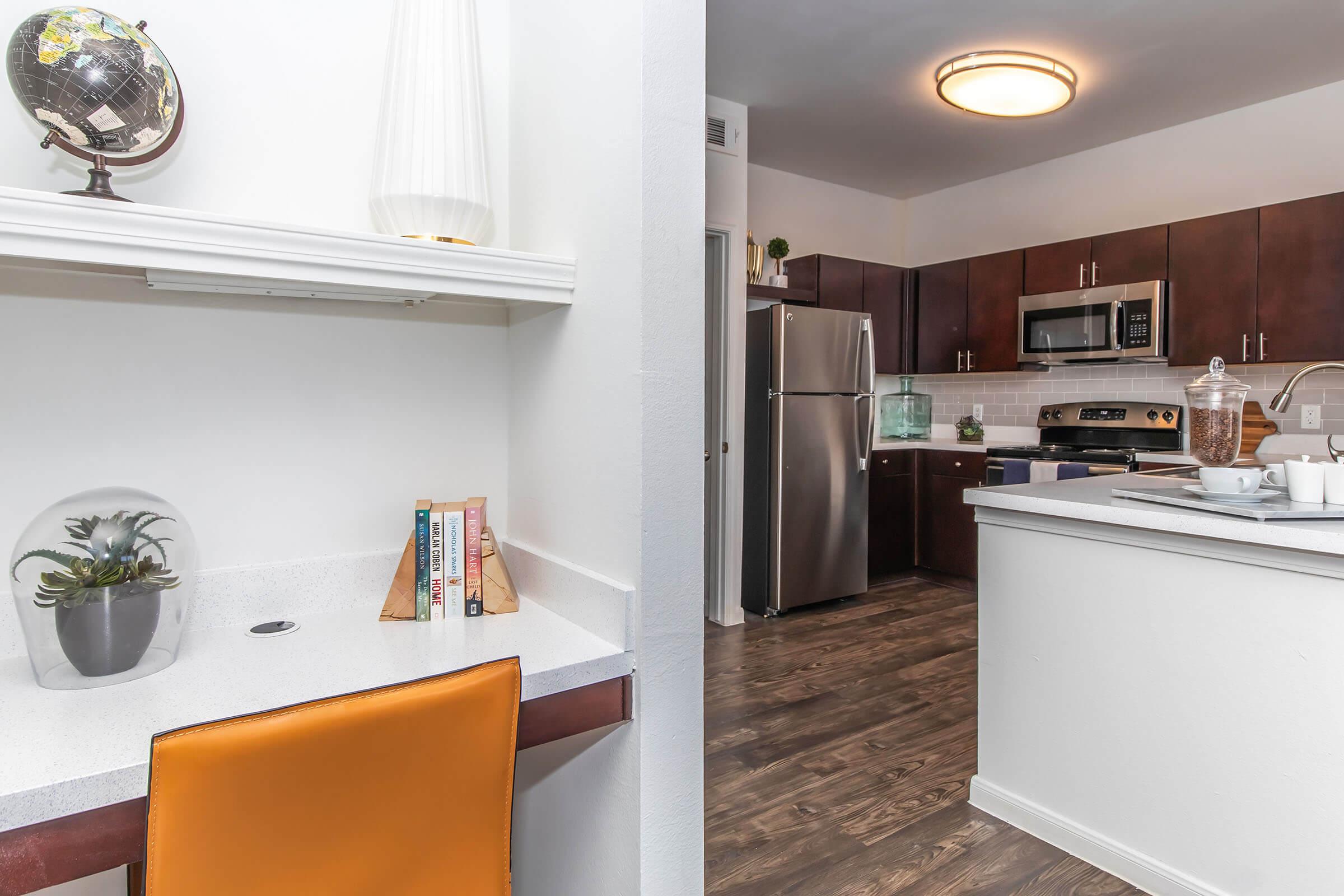 A modern kitchen featuring dark wood cabinets, stainless steel appliances, and a quartz countertop. In the foreground, a small workspace with an orange chair, a potted plant, and books arranged on a shelf. A globe on a stand is positioned nearby, adding a decorative touch to the room's contemporary design.