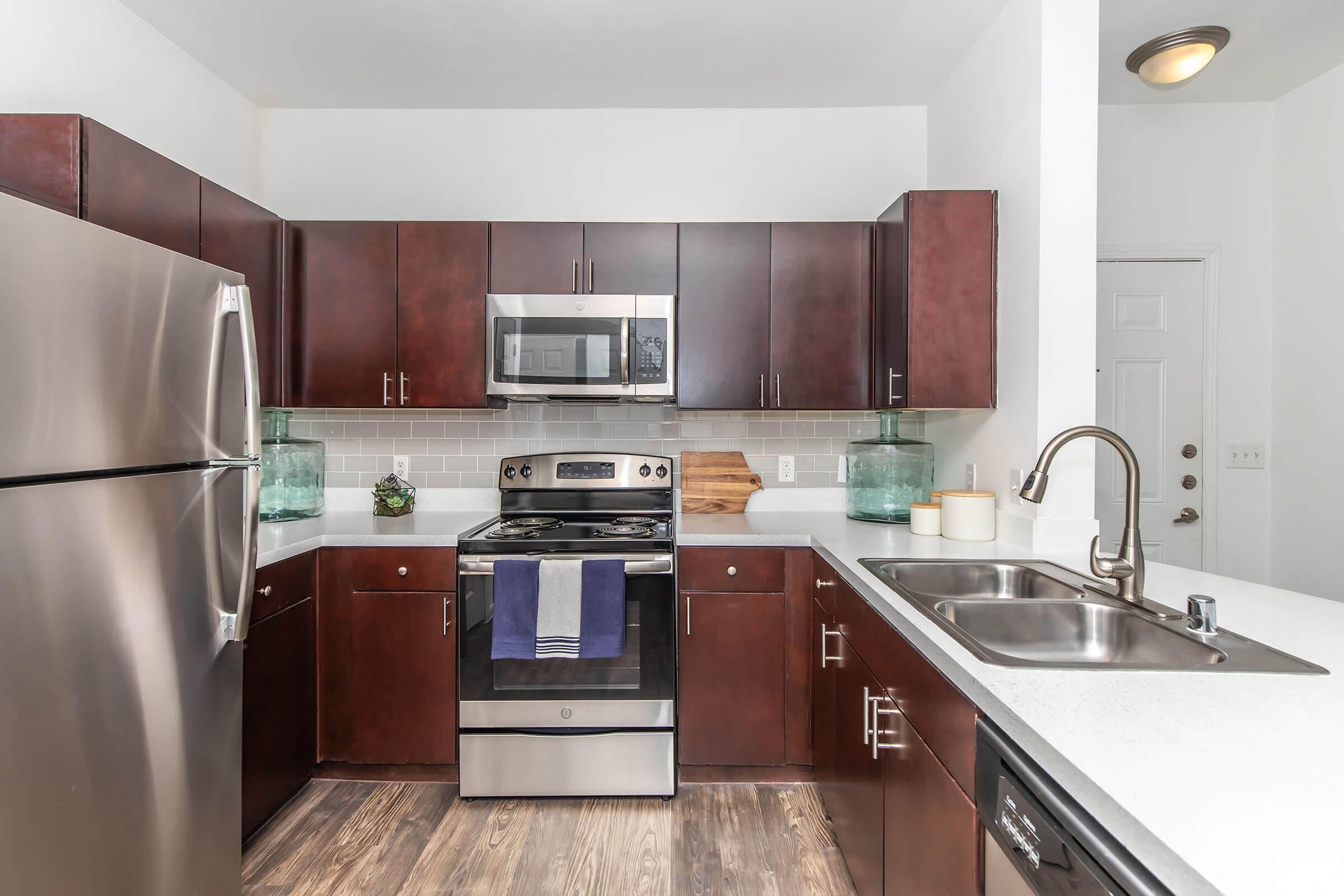 Modern kitchen featuring dark wood cabinets, stainless steel appliances, a double sink, and a gray tiled backsplash. The countertop is light, and there's a small decorative plant and glass containers on display. The space is well-lit and organized, with a door visible leading to another area.
