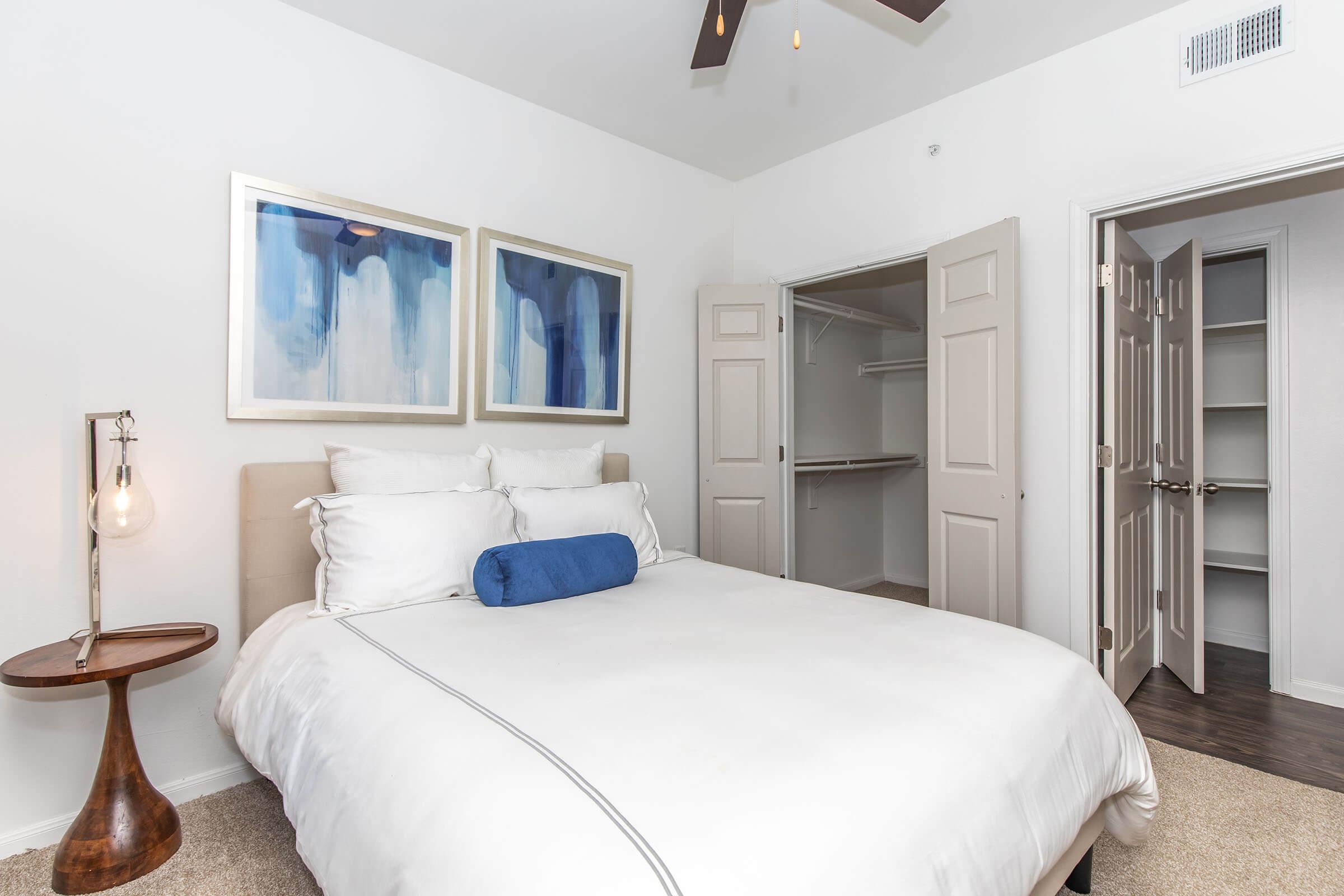 A bright, modern bedroom featuring a queen-sized bed with white linens and a blue decorative pillow. Two framed abstract artworks hang above the bed. A wooden side table with a pendant lamp is beside the bed. An open closet is visible in the background, showcasing sliding doors and a tidy space.