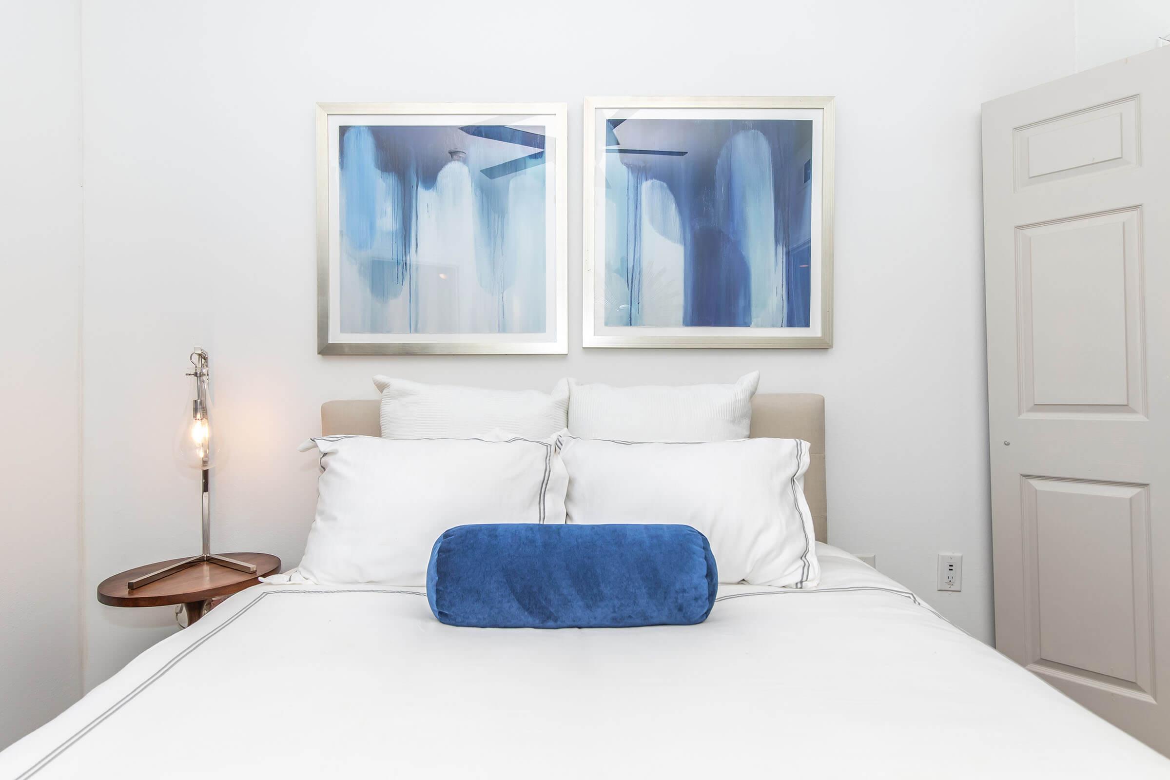 A cozy bedroom featuring a neatly made bed with white pillows and a blue bolster pillow in the center. Above the bed are two framed abstract paintings in shades of blue. A small, modern bedside table is visible, along with a stylish light fixture. The walls are painted white, creating a fresh atmosphere.