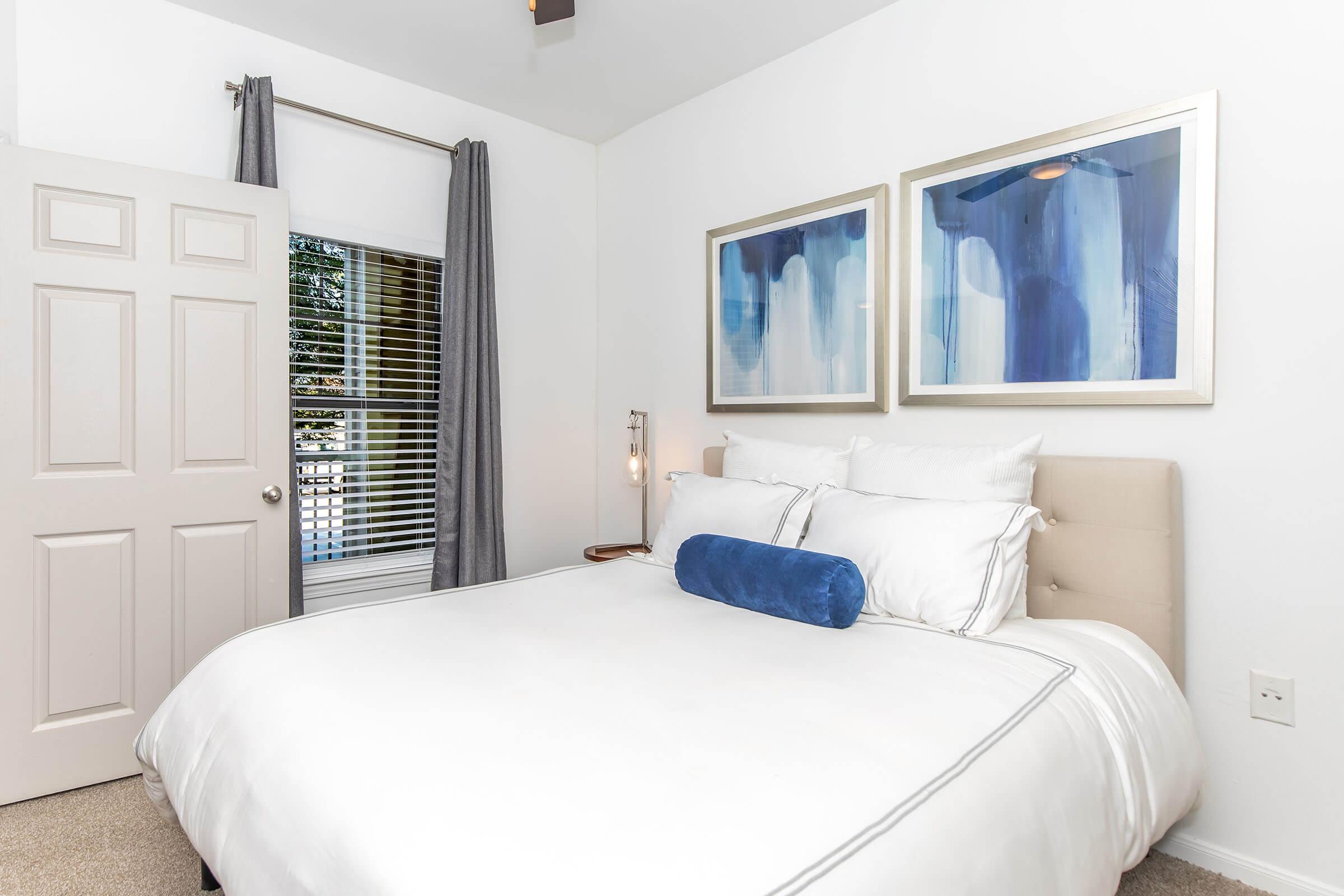 A cozy bedroom featuring a queen-sized bed with white linens and a blue bolster pillow. Two abstract blue art pieces are framed on the wall. A window with white blinds allows natural light, and gray curtains add a touch of elegance. A closed door is visible, completing the inviting atmosphere.