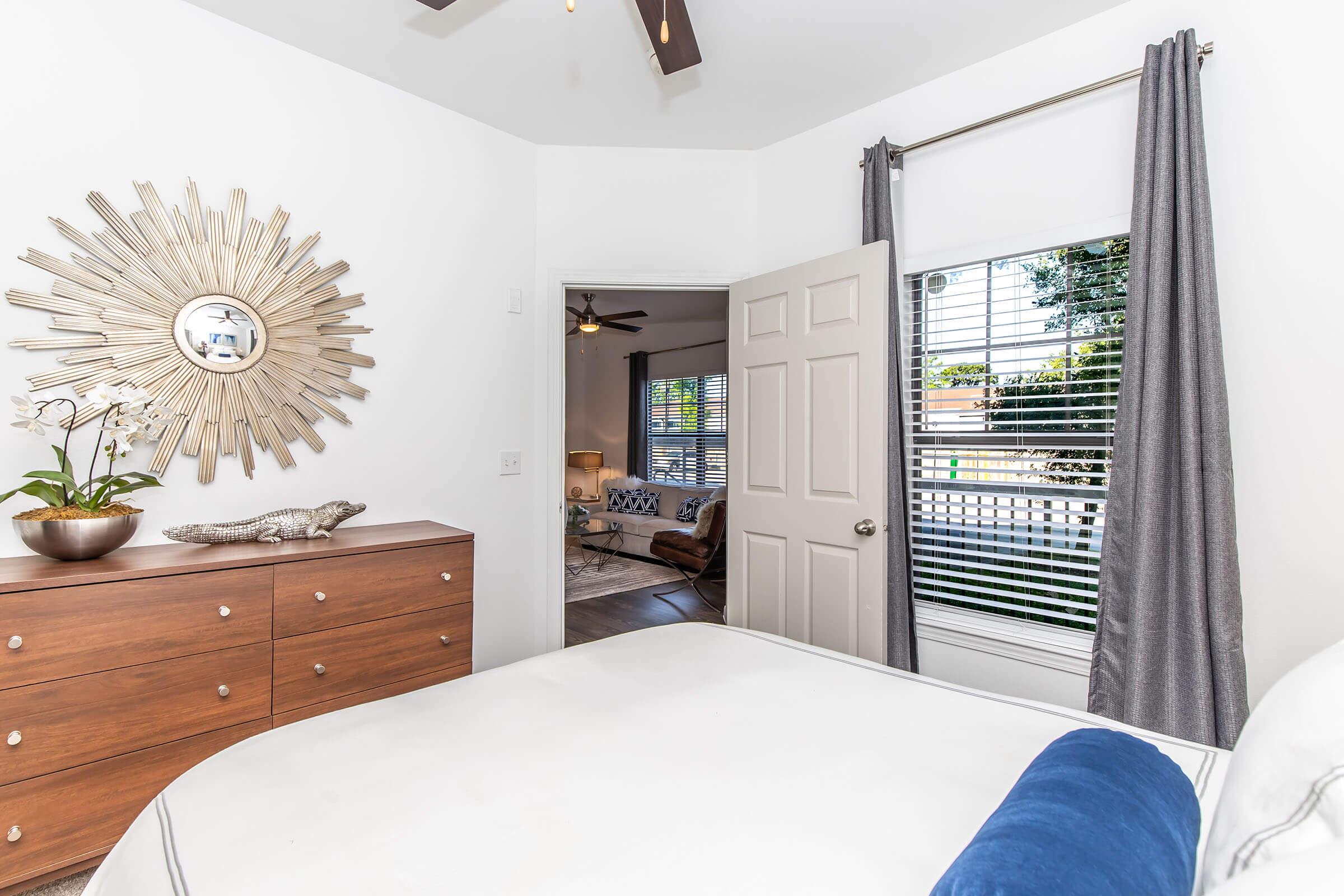 A cozy bedroom featuring a wooden dresser with decorative items, a sunburst mirror on the wall, and an open door leading to a living area. The room is well-lit with natural light streaming through a window adorned with gray curtains, and a plush blue throw pillow on the bed adds a pop of color.