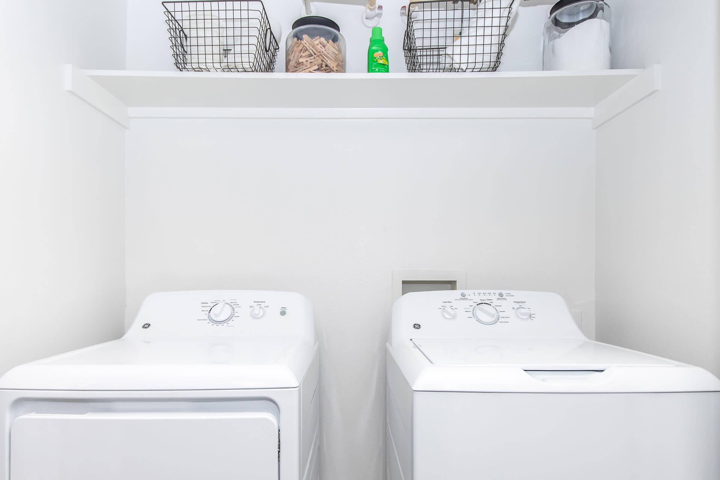 A clean and bright laundry room featuring a stacked washer and dryer, both in white. Above them, a shelf holds baskets and a bottle of detergent, adding organization and efficiency to the space. The walls are painted white, contributing to a fresh and modern look.