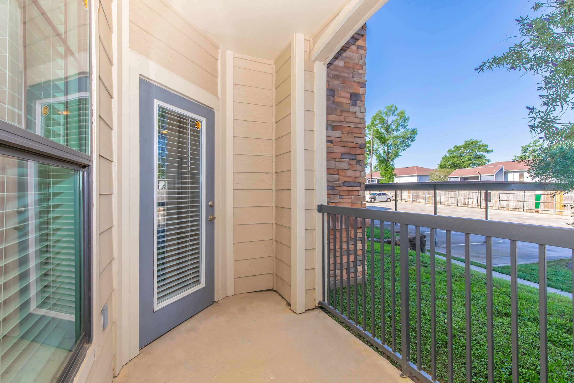 A small covered balcony with a gray door featuring vertical blinds. The railing has a simple design, and there is a view of greenery and a parking lot beyond. The walls are light-colored siding with a stone accent on one side. Bright, sunny day with clear blue sky.
