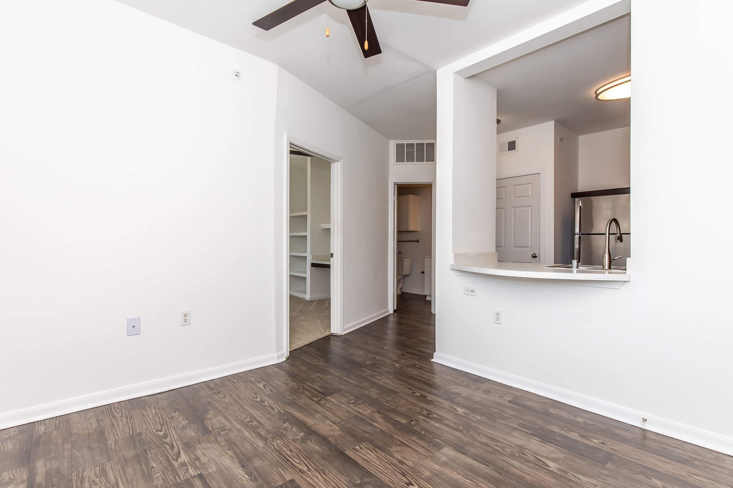 Interior view of a bright, modern living space featuring light-colored walls, dark wood flooring, and a ceiling fan. A doorway leads to another room, and an open kitchen area with a bar seating counter is visible. The space has natural light and a clean, minimalist design.