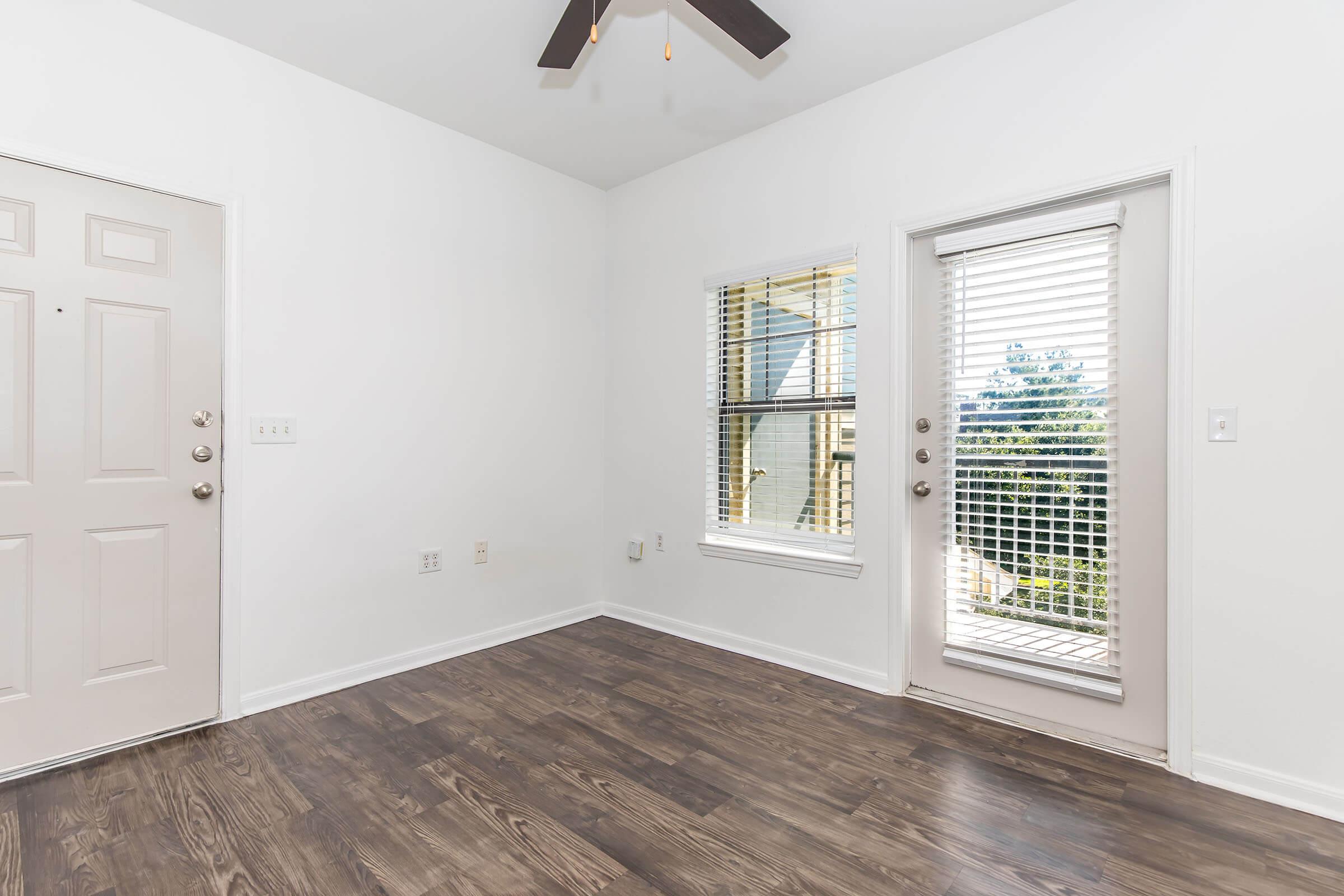 A bright and spacious room featuring a light-colored wall, a ceiling fan, and a large window with blinds. There is a door leading outside and a standard entry door on the left. The floor is covered with dark wooden laminate, creating a warm and inviting atmosphere.