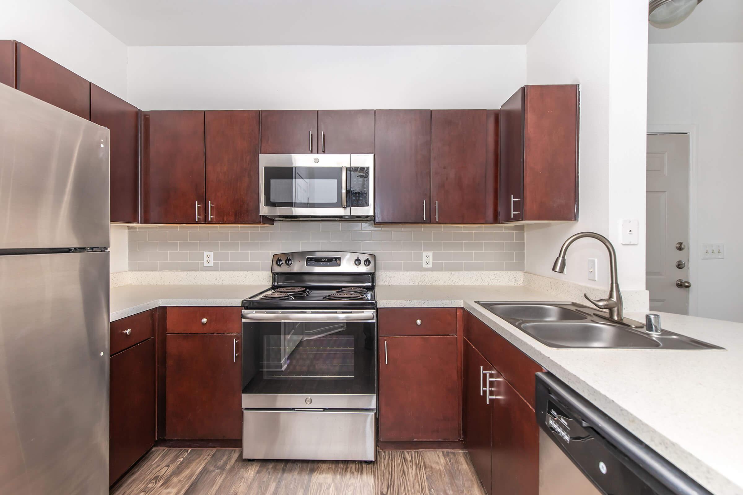 A modern kitchen with dark wooden cabinets, stainless steel appliances, including a refrigerator and stove, and a microwave mounted above the stove. The countertop is light-colored with a sink and faucet, and the walls are painted white, creating a clean and bright atmosphere.